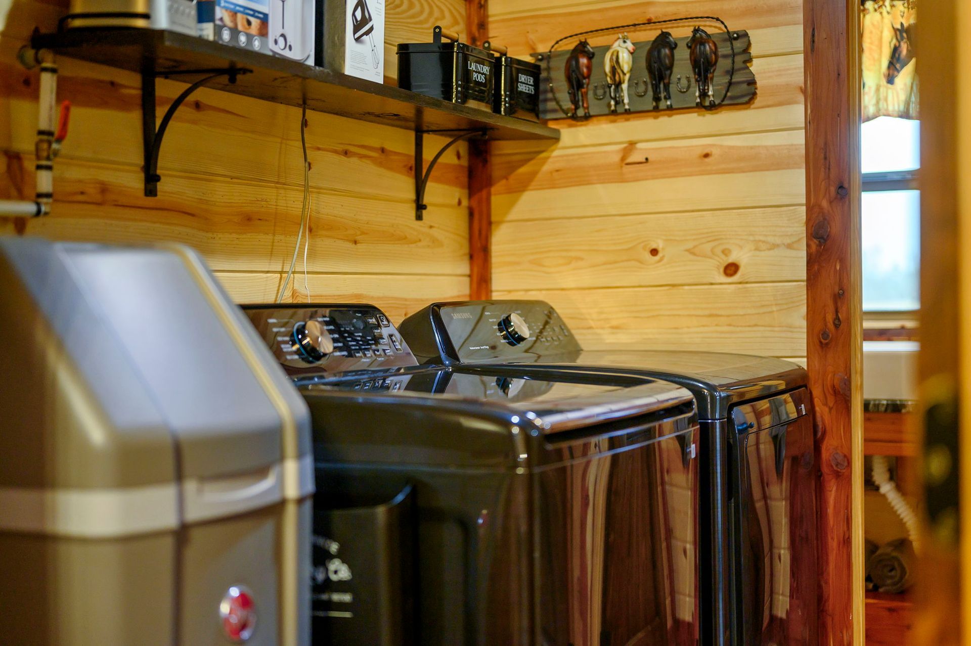 A laundry room with dark gray appliances, a water softener, and a shelf with decorative items against wood paneled walls.