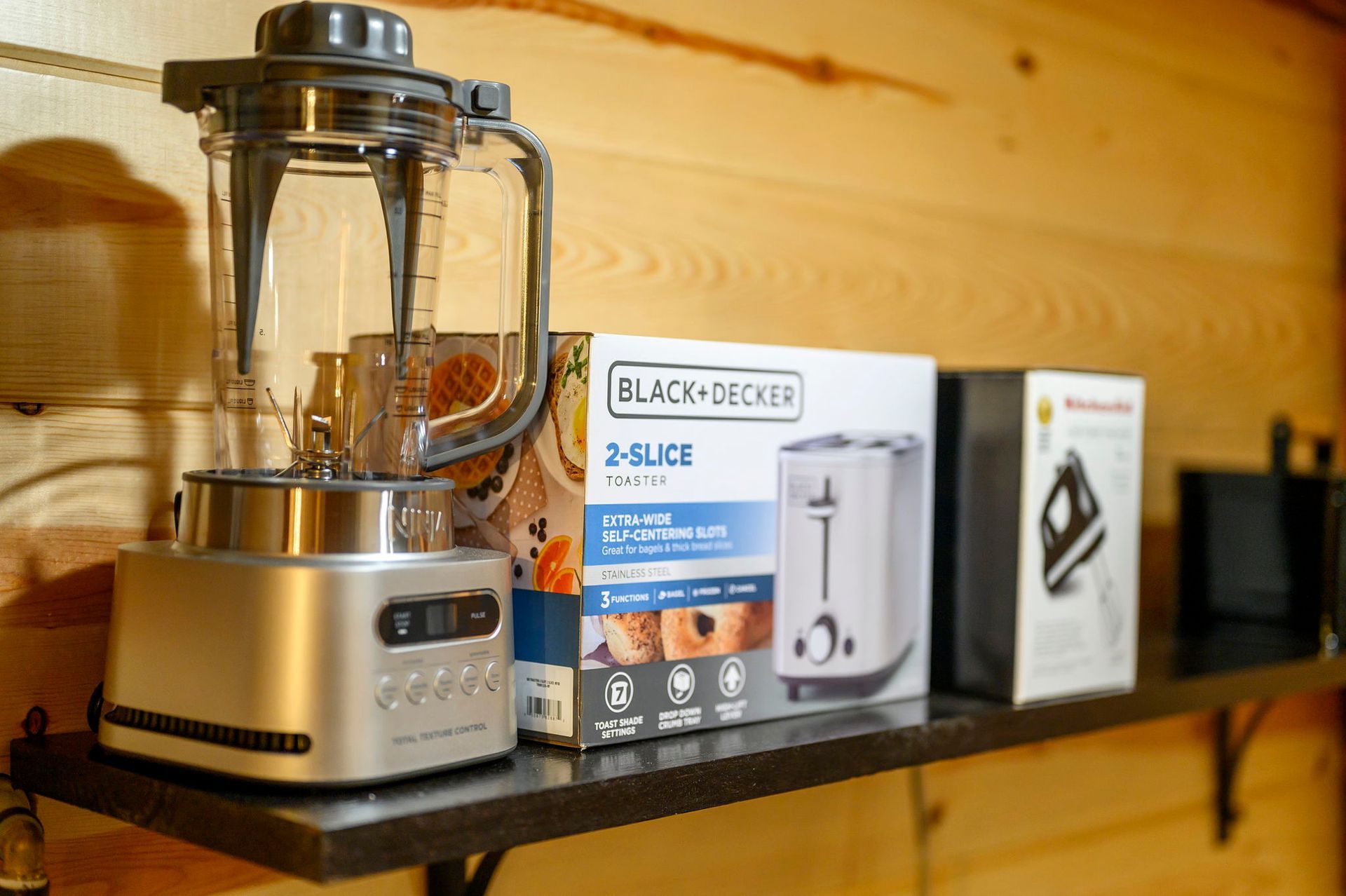A silver blender, a Black+Decker toaster box, and a smaller appliance box sit on a dark shelf against a wood-paneled wall.