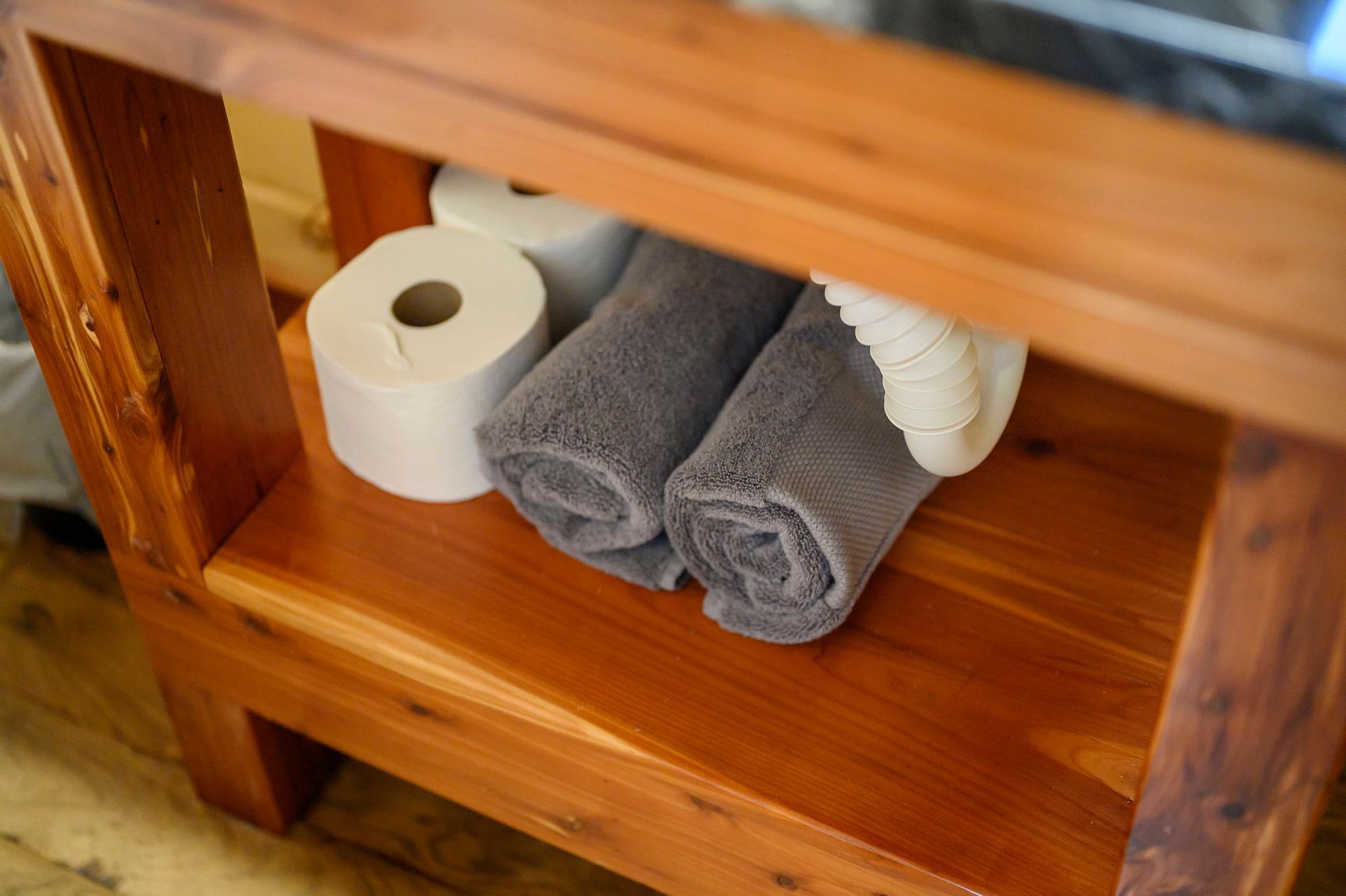A wooden vanity shelf holds two rolls of toilet paper, two rolled-up gray towels, and a white pleated fabric item.
