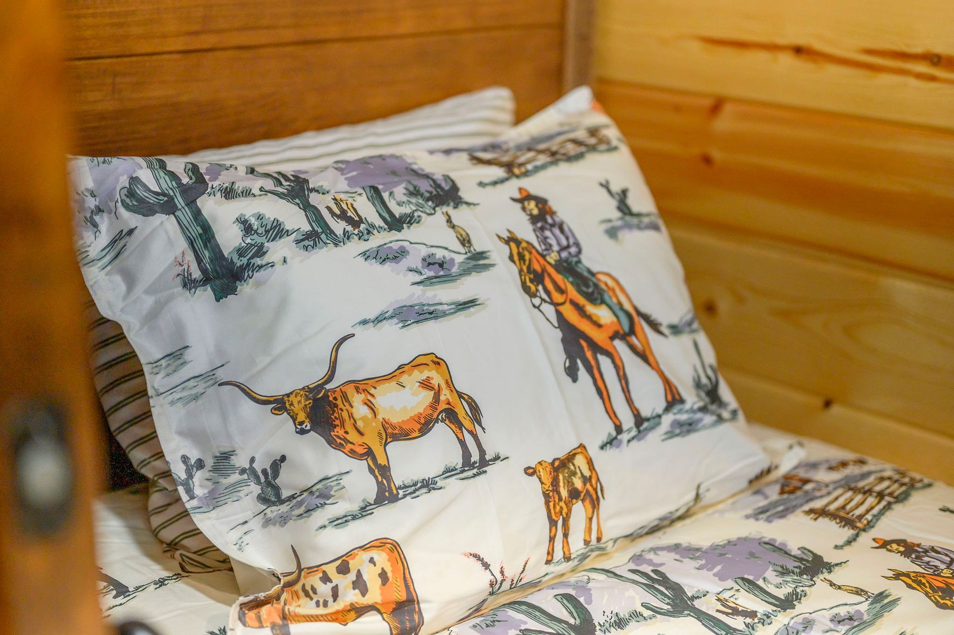 A decorative pillow with a Western-themed print of a cowboy on a horse and longhorn cattle on a bed with wood walls.
