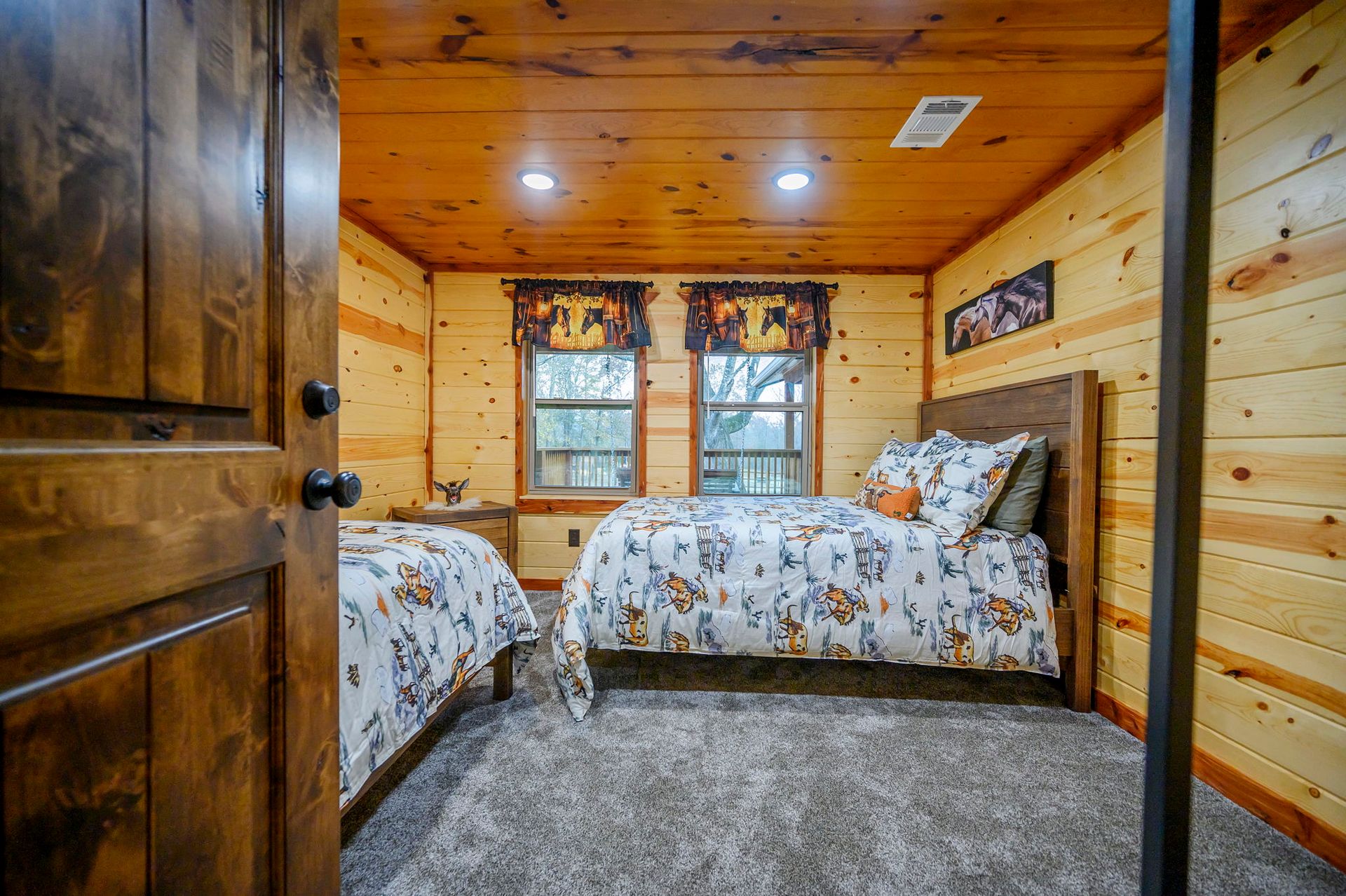 A rustic bedroom with wood-paneled walls, two beds with patterned bedding, grey carpet, and two windows with curtains.