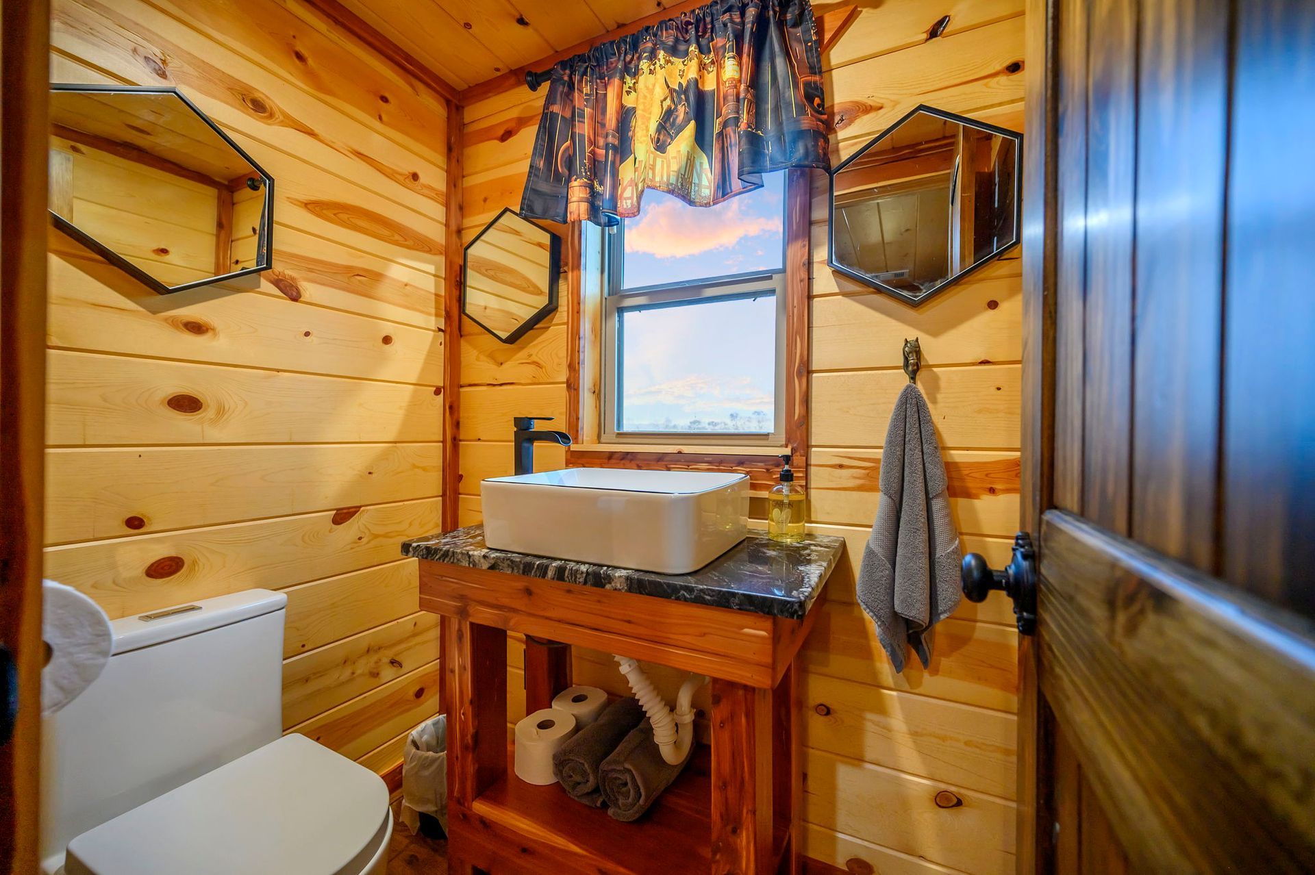 A small rustic bathroom featuring wooden walls, a white sink on a wood vanity, a toilet, two wall mirrors, and a window.