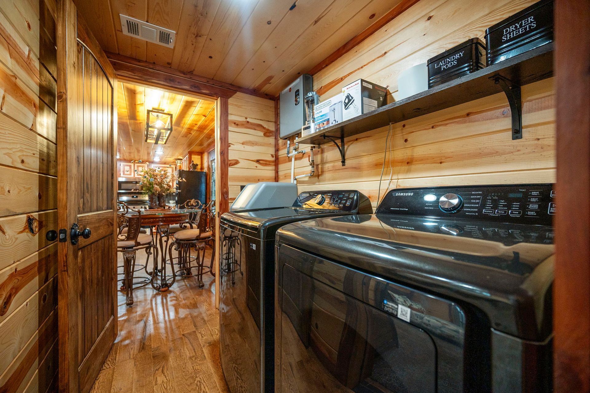 A laundry room with dark appliances and wood-paneled walls, looking through a doorway into a furnished dining area.