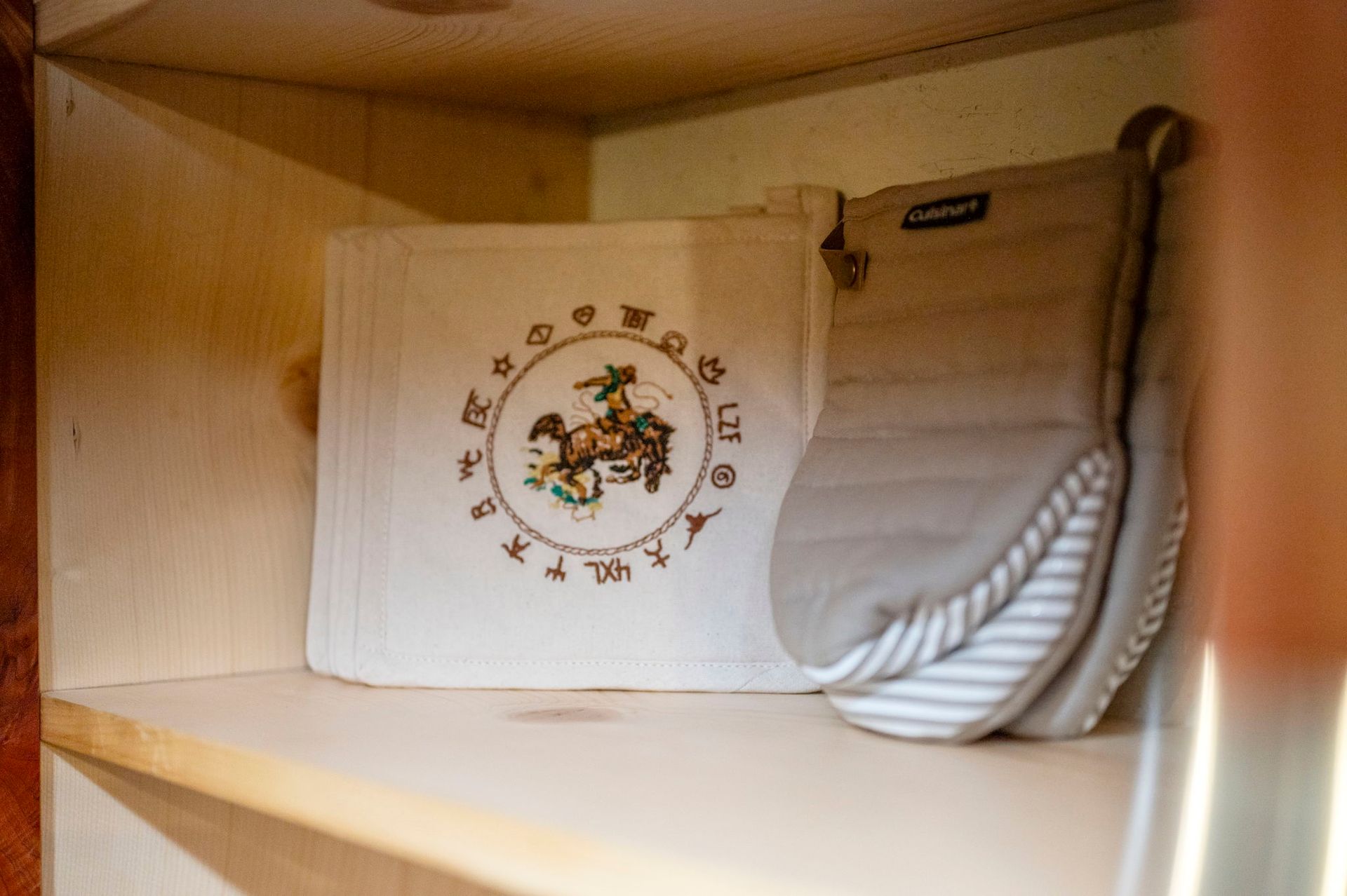 A white cloth embroidered with a circular design sits next to a folded gray and white striped oven mitt on a wood shelf.