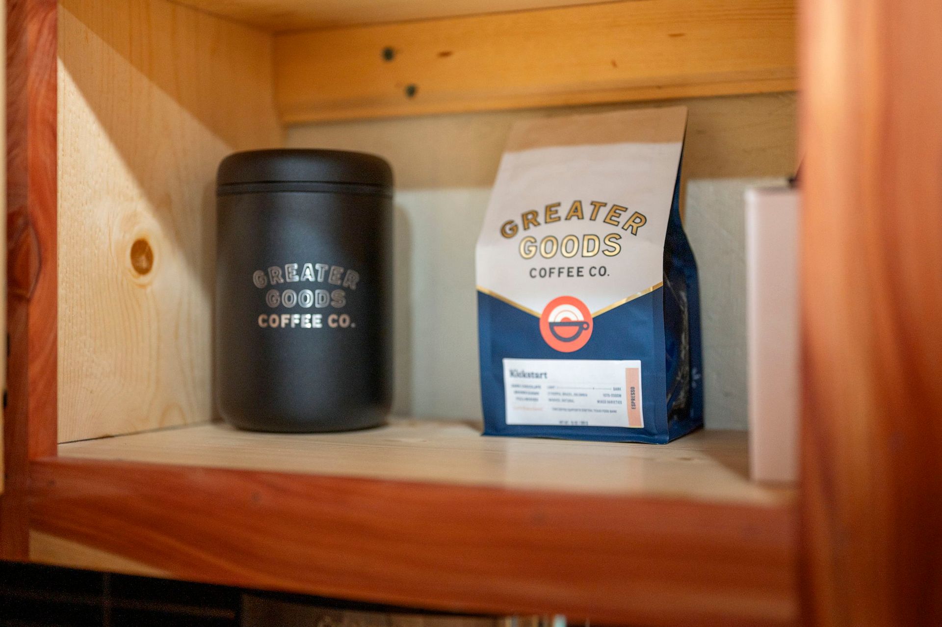 A black canister and a bag of Greater Goods Coffee Co. beans sitting on a wooden shelf inside a cabinet.