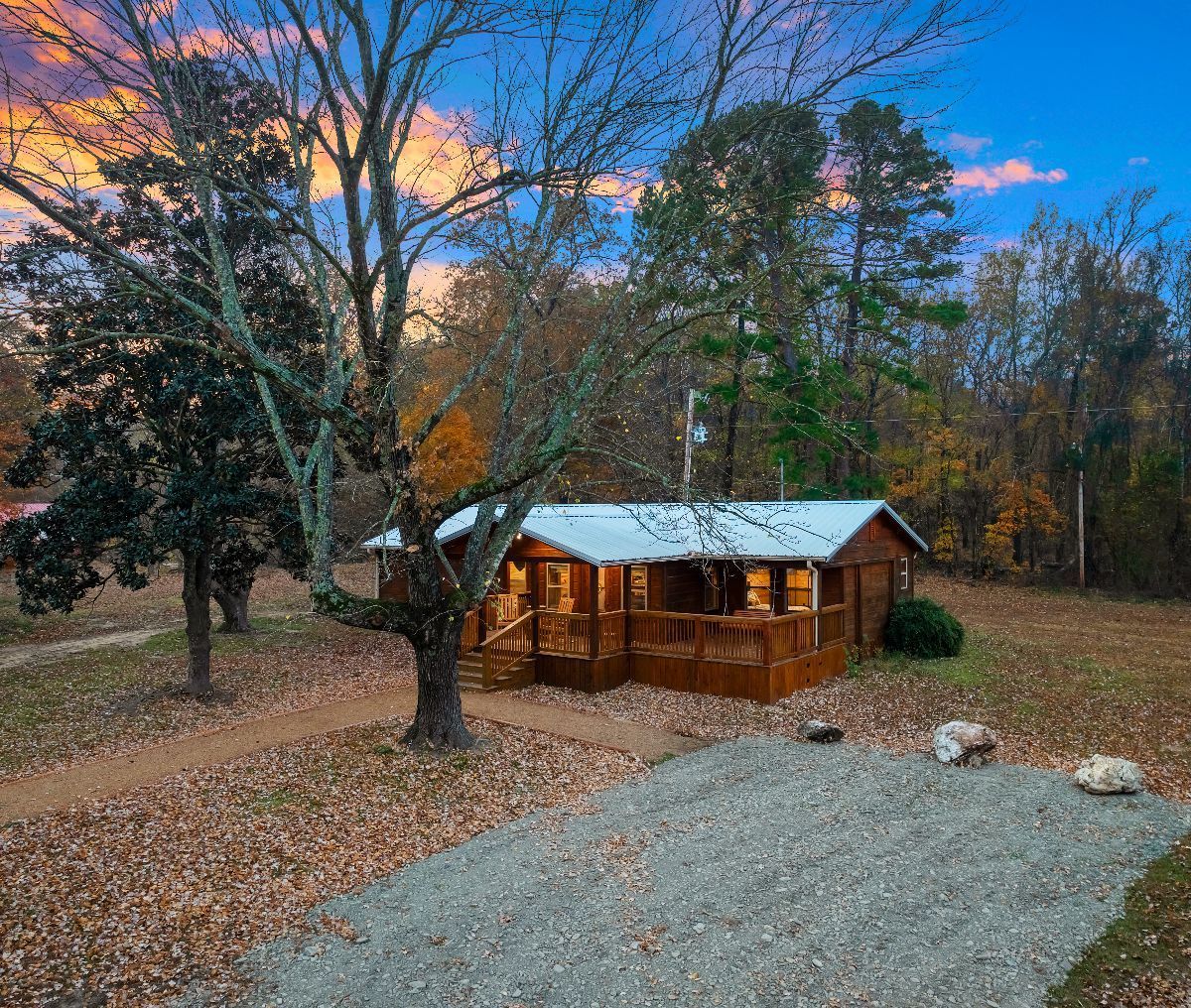 A rustic wooden cabin with a metal roof and a wrap-around porch sits in a wooded yard under a sunset sky.