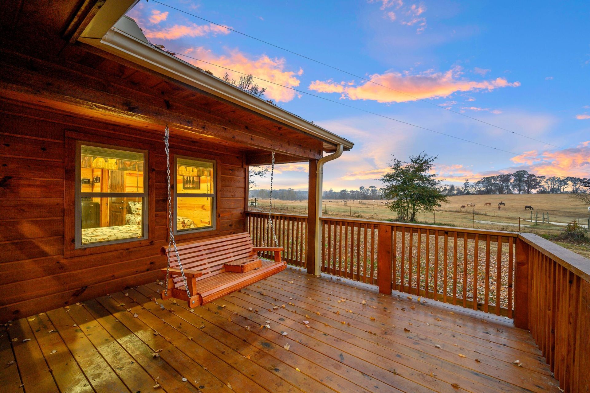 Wooden porch with a hanging bench, overlooking a vast, grassy field under a vibrant sunset sky.