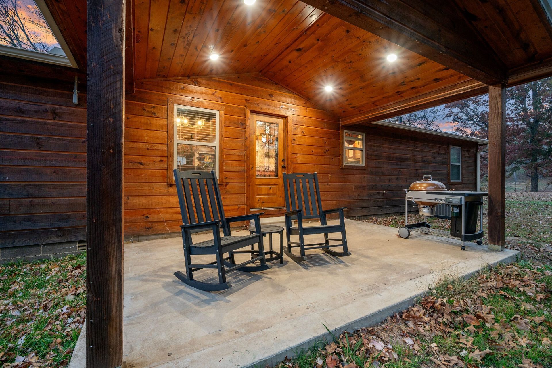 Two black rocking chairs sit on a covered concrete patio outside a rustic wooden cabin at twilight.