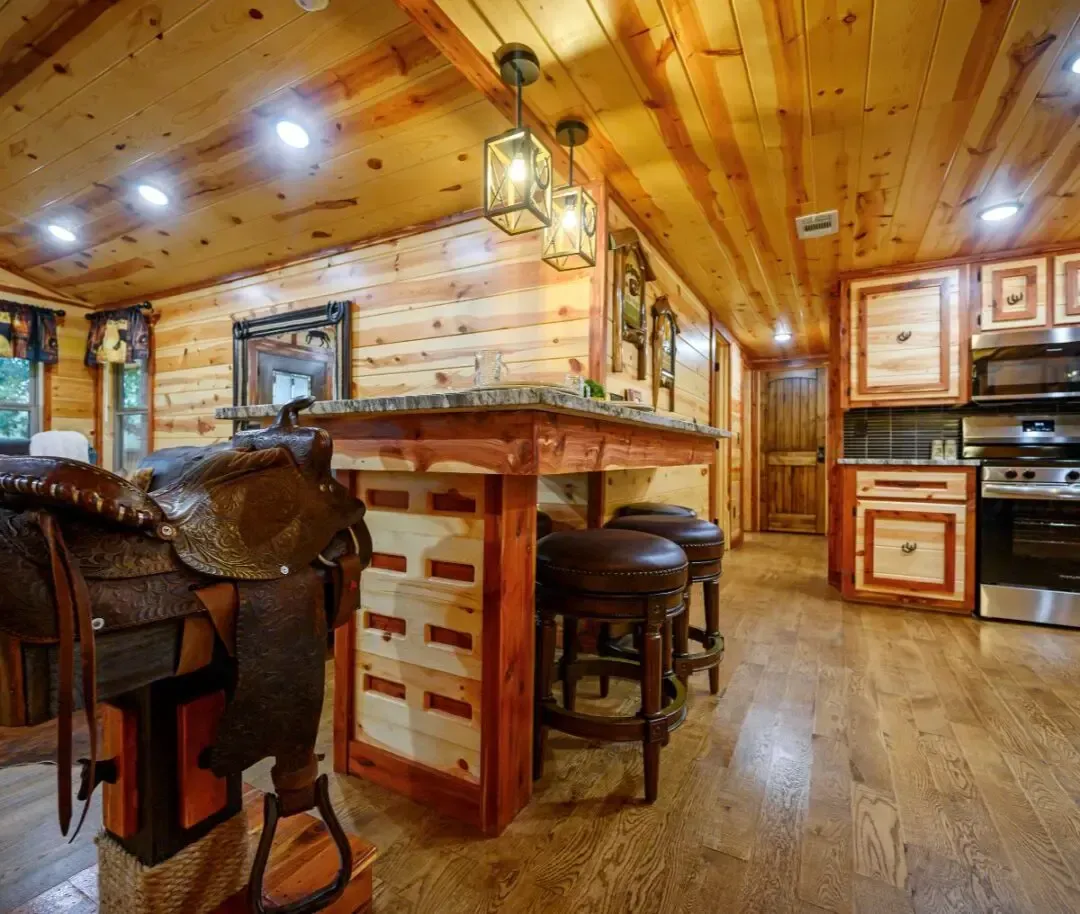A rustic kitchen with wood-paneled walls and ceilings, a wooden island with bar stools, and a leather saddle on display.
