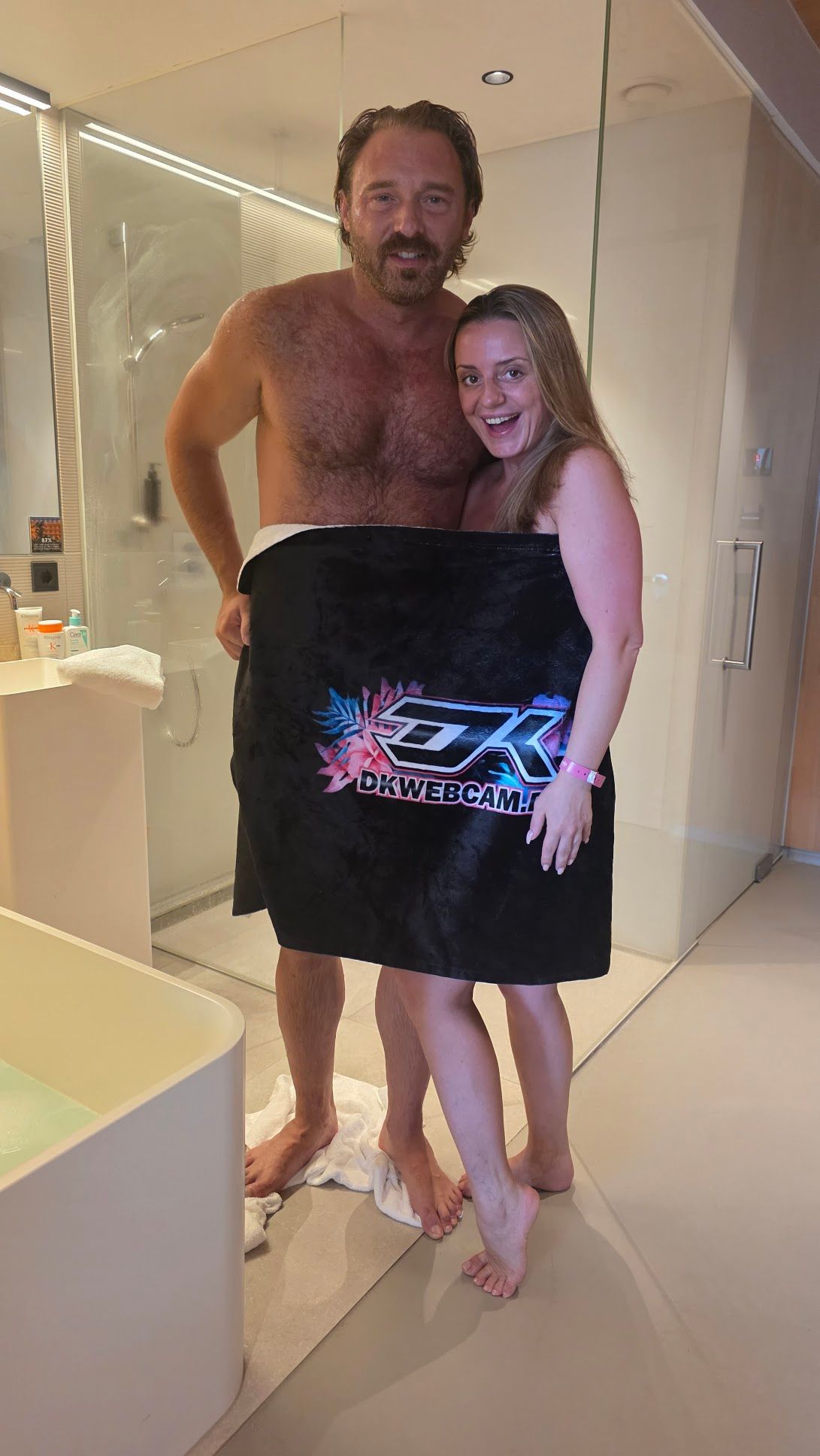 Man and woman wrapped in a towel, smiling, standing in a modern bathroom, shower in background.