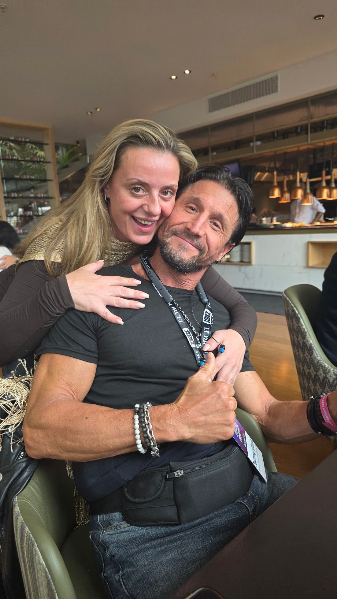 Woman hugs man indoors, smiling. Man in black shirt, thumbs-up. Restaurant setting.