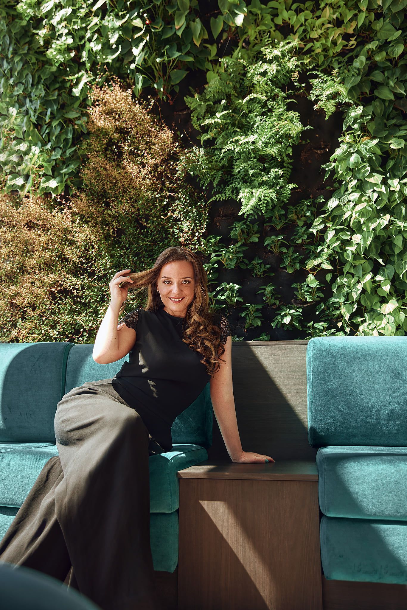 Woman seated on a teal couch, smiling, touching her hair; green wall in the background.