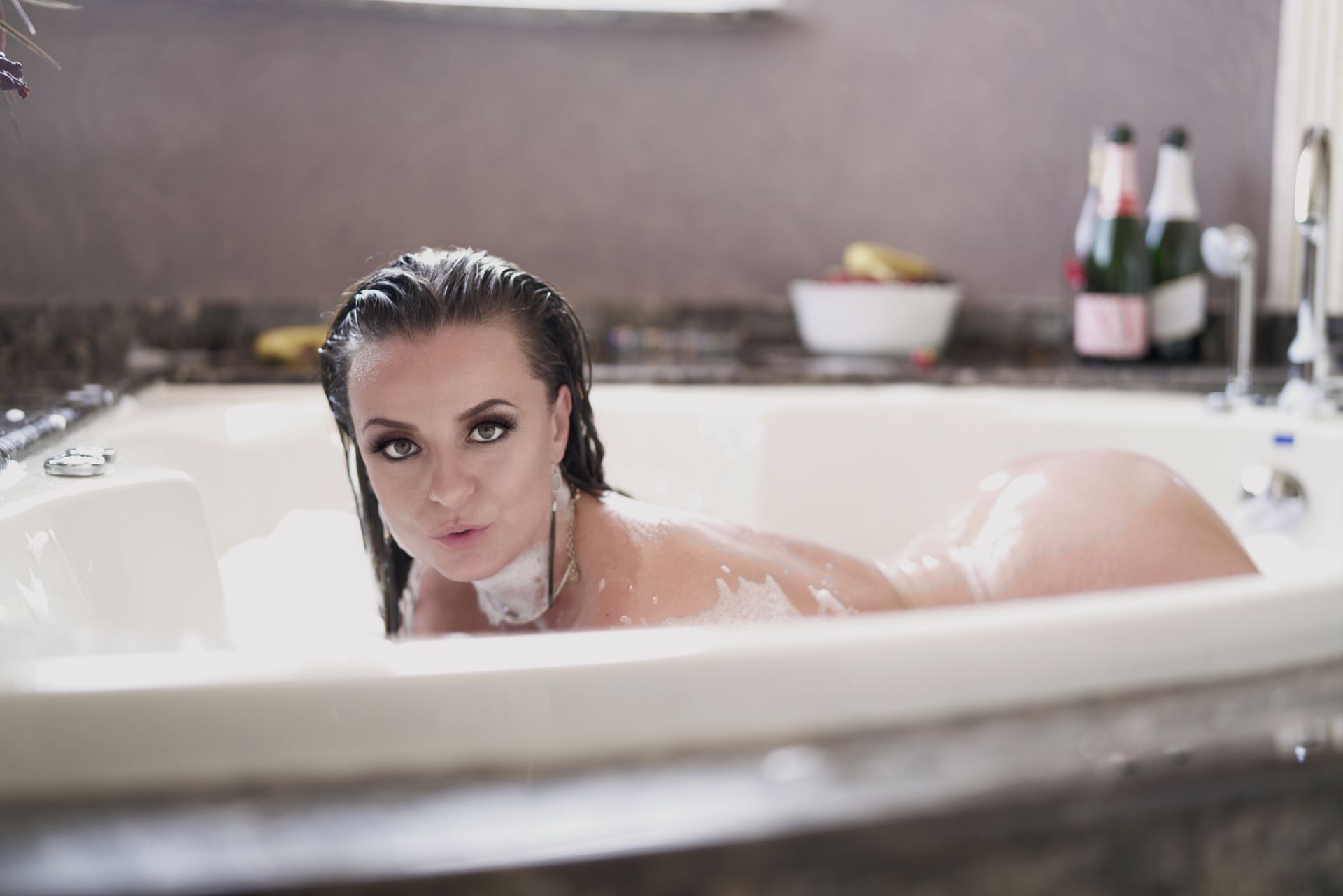 Woman in bathtub blowing kiss, wet hair, bubbles, champagne bottles in background.