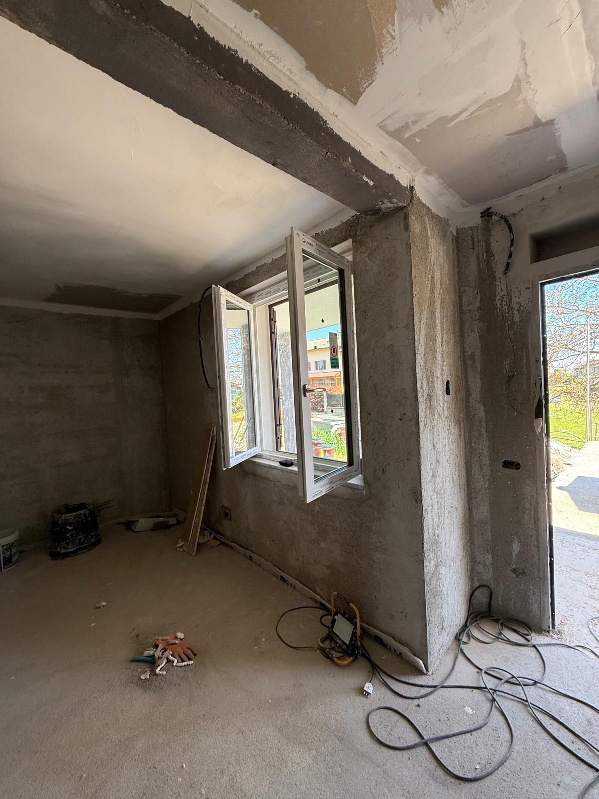 Interno di una stanza in costruzione con pareti in cemento a vista, finestre e cavi elettrici.