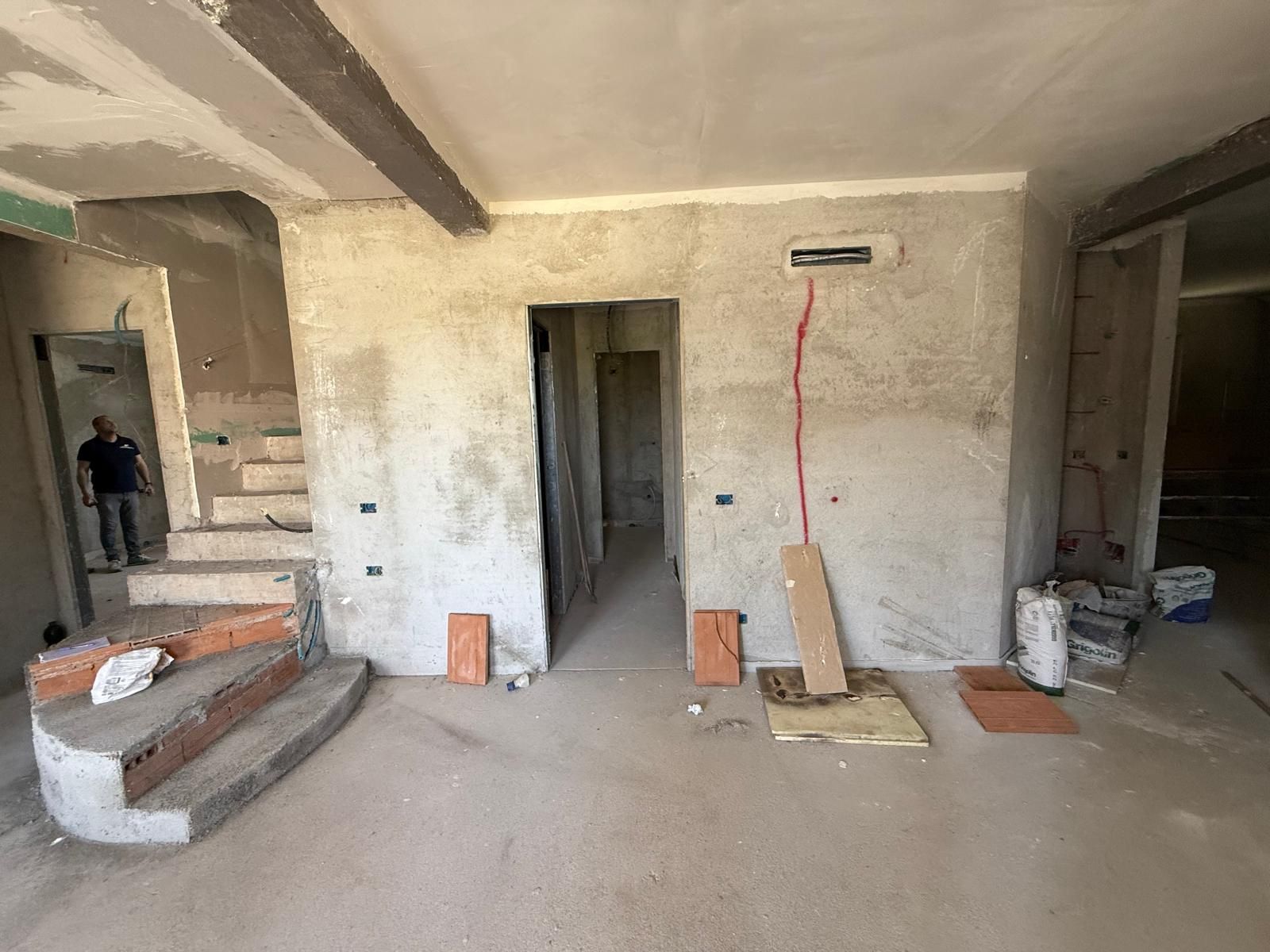 Interno di un edificio in costruzione. Un uomo è in piedi vicino a una scala con mattoni a vista. Sono visibili pareti grigie e pavimenti grezzi Geotecno Genova