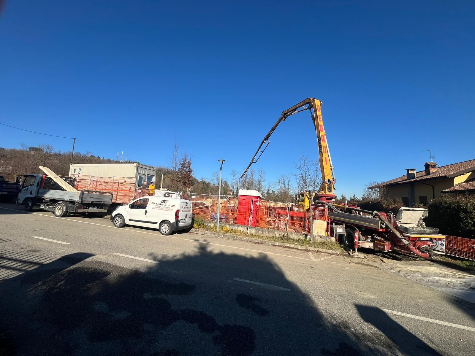 Cantiere edile con un'autopompa per calcestruzzo che estende il suo braccio, un furgone bianco e altri veicoli parcheggiati su una strada asfaltata contro un cielo azzurro.