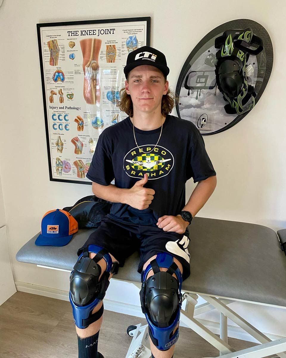 A Young Man With Knee Braces is Sitting on a Table Giving a Thumbs Up — Gall Bros Medical in Robina, QLD