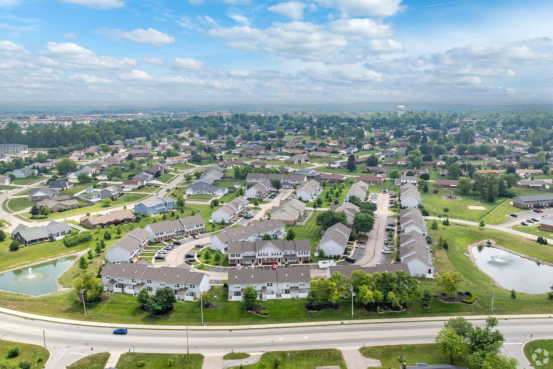 Aerial view of a suburban neighborhood with houses, roads, ponds, and greenery under a blue sky.