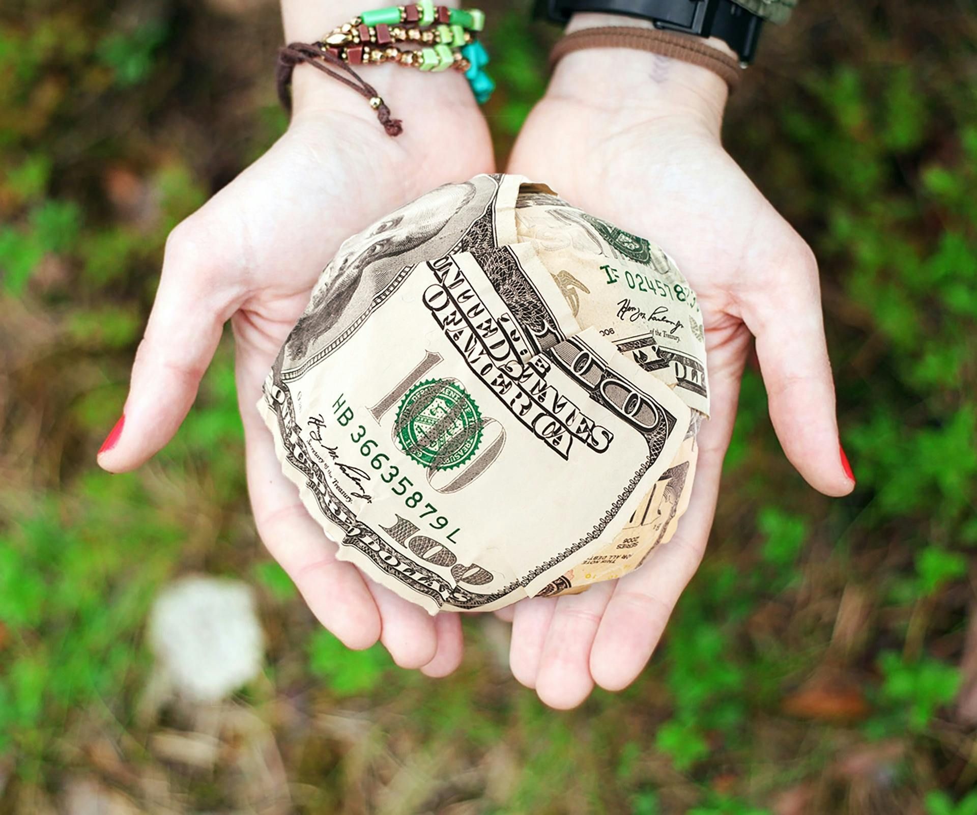 Hands holding a round paper-cutout with a dollar bill design over a blurred green outdoor background
