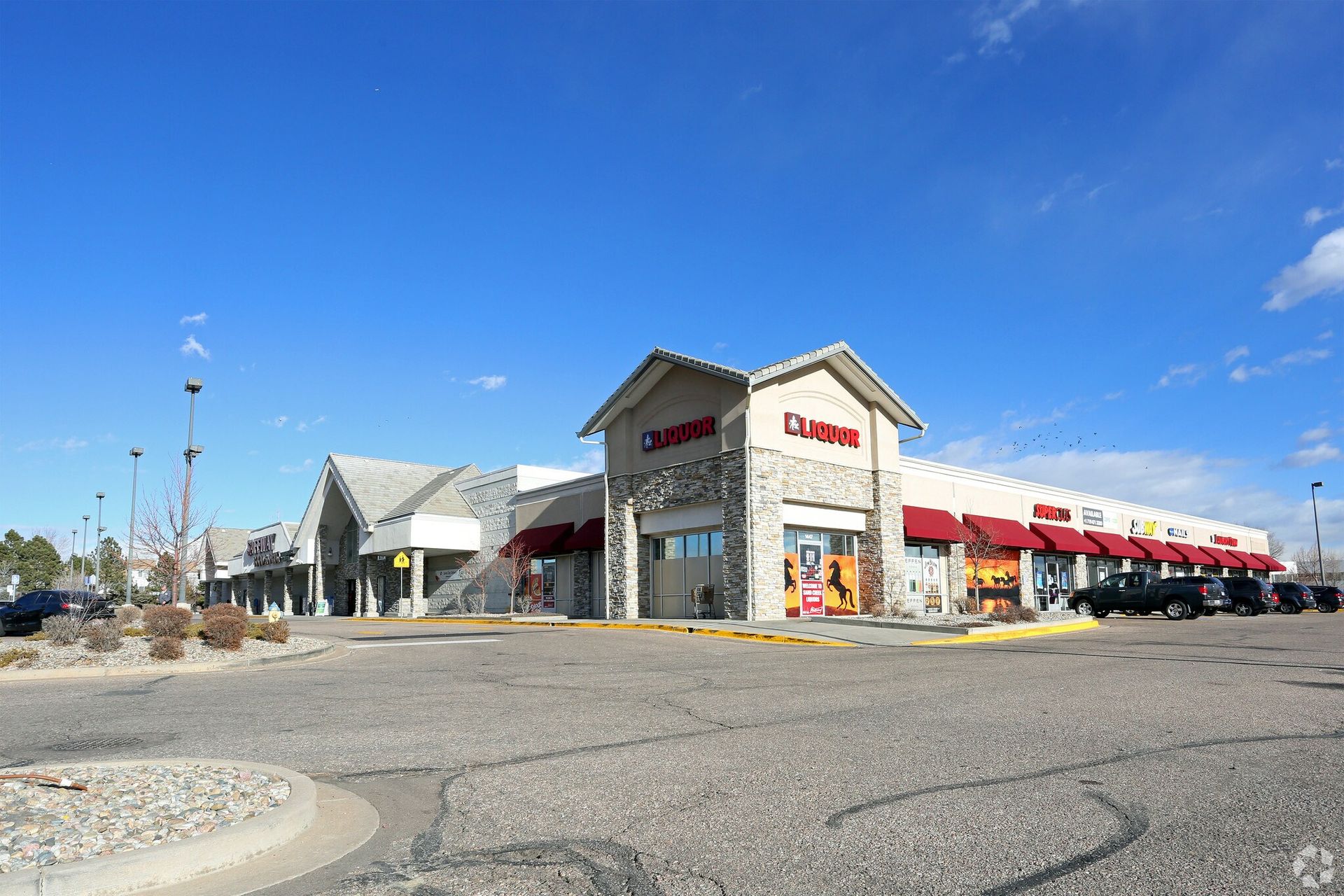 Exterior view of a commercial building with multiple storefronts, stone and red accents, under a clear blue sky.