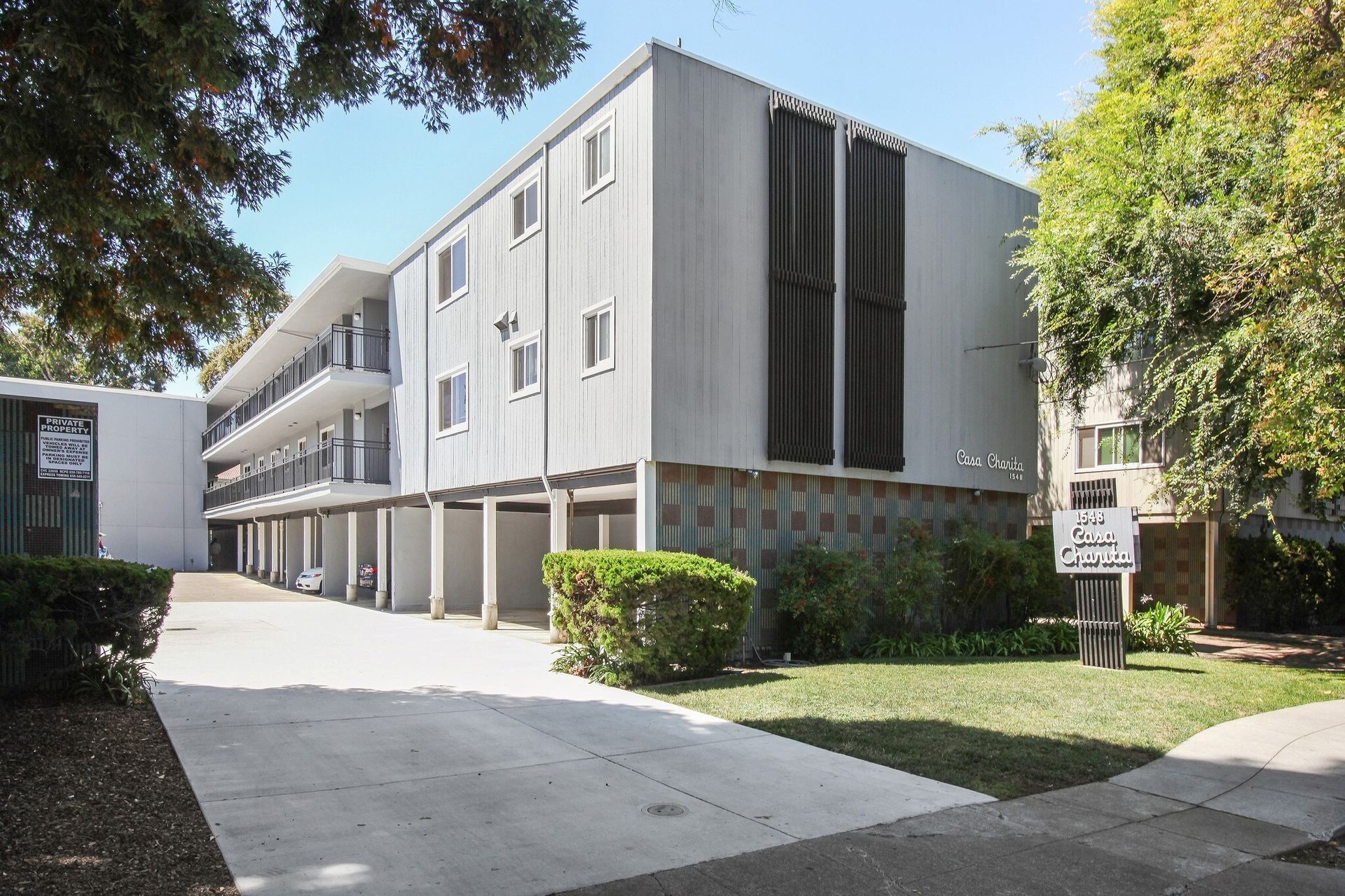 Three-story gray apartment building with balconies, supported by pillars, on a grassy lot.