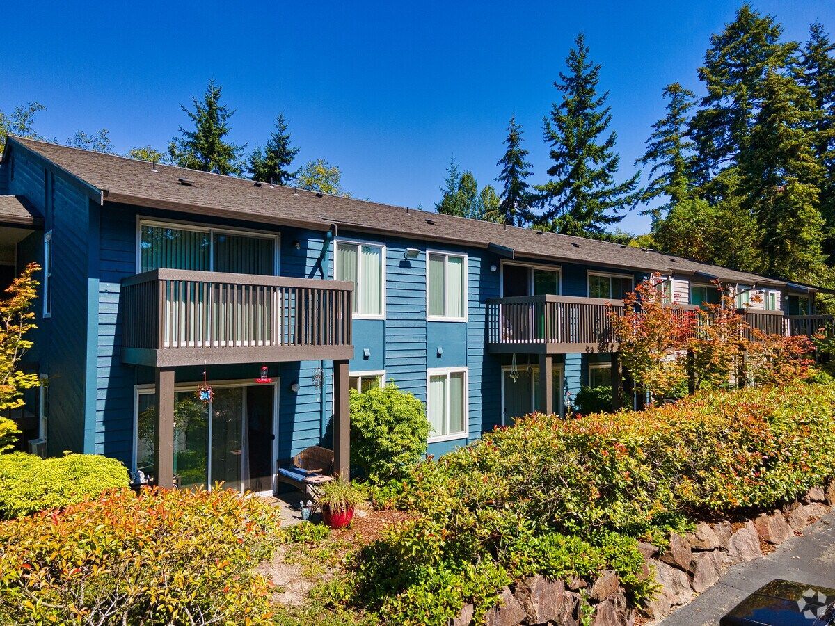 Blue apartment building with balconies, surrounded by bushes and trees under a clear sky.