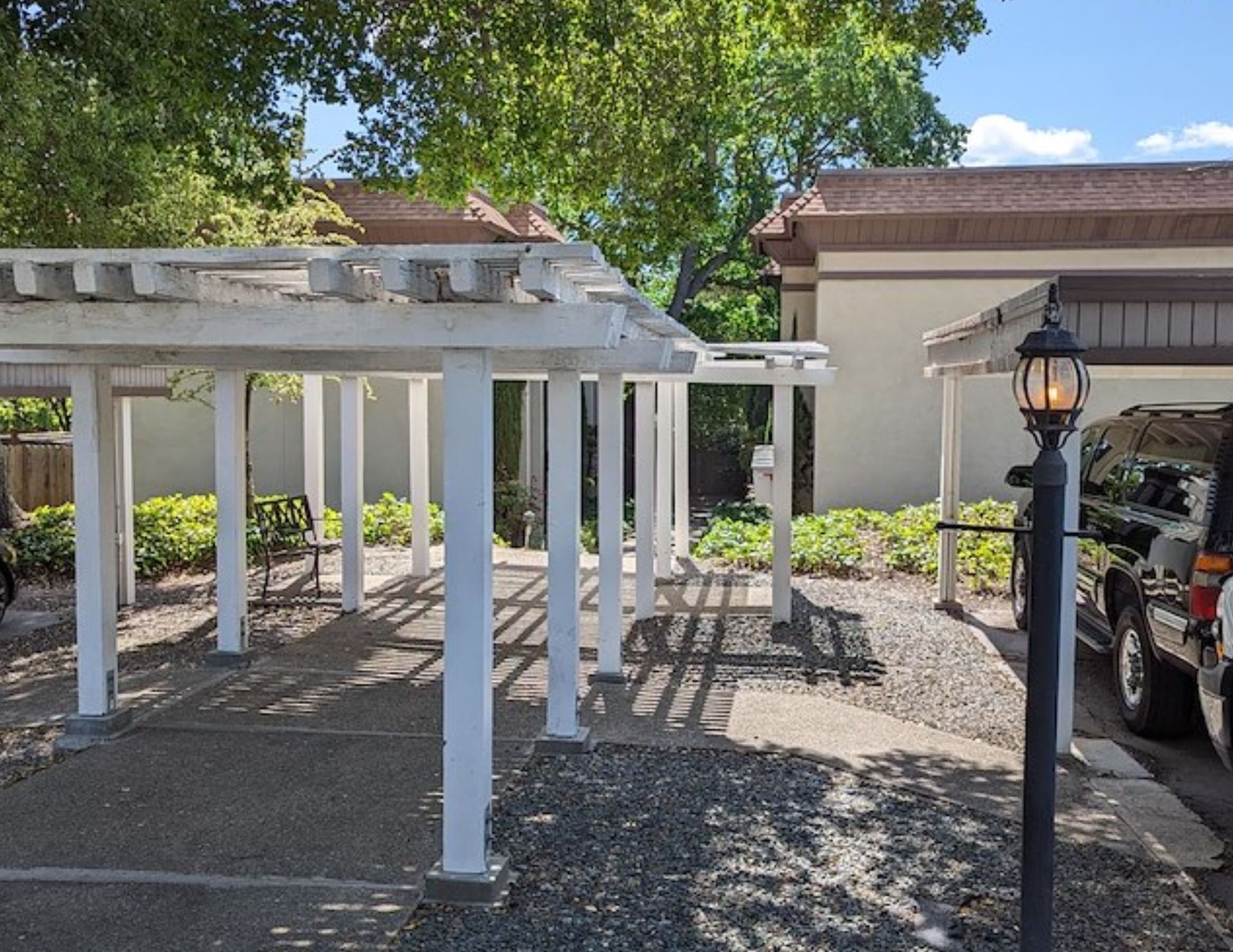 White pergola over a parking area, with a lamppost on the right. Buildings and trees are in the background.