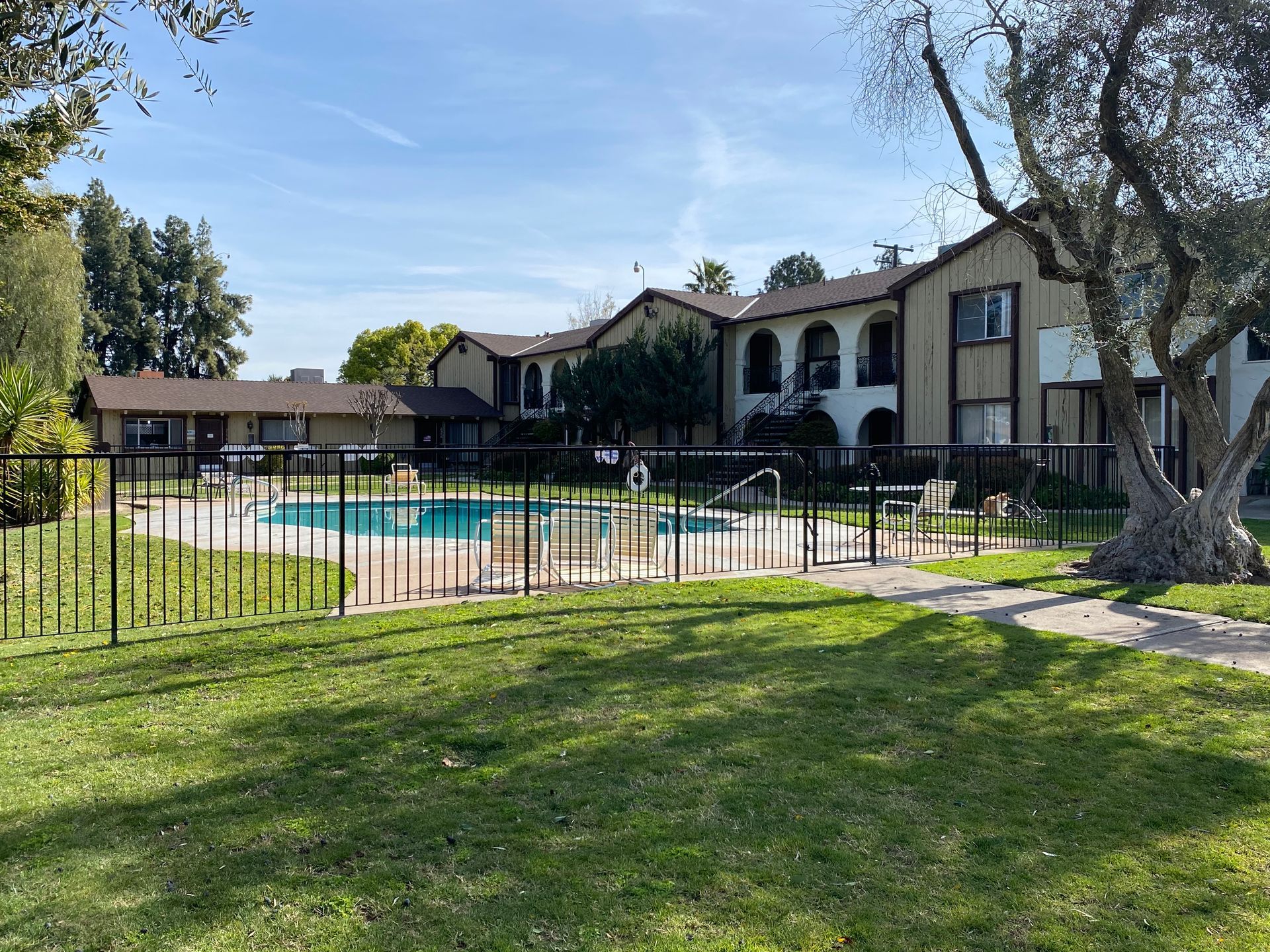 Apartment complex with a fenced swimming pool and green lawn on a sunny day.