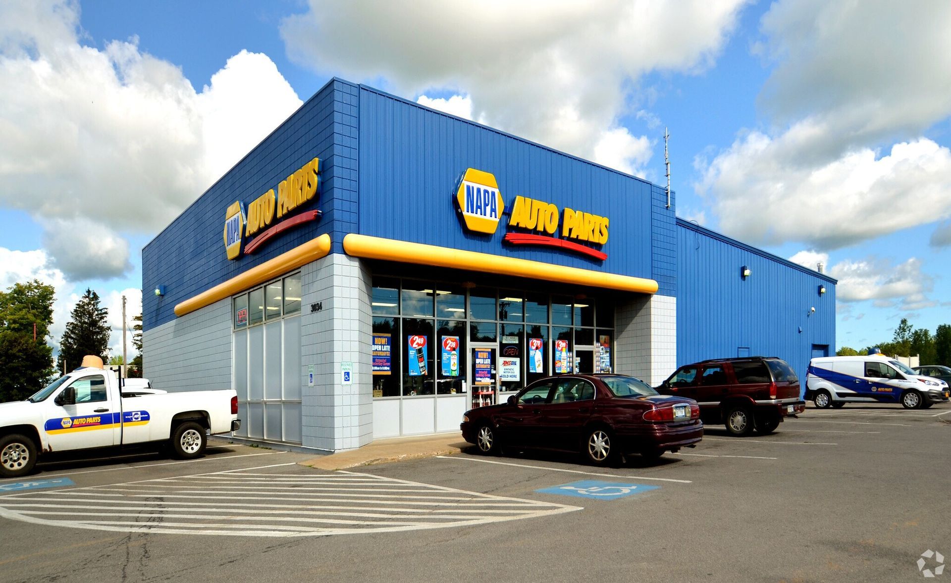 NAPA Auto Parts store exterior, blue building with yellow trim, parked cars and a delivery truck on a sunny day.
