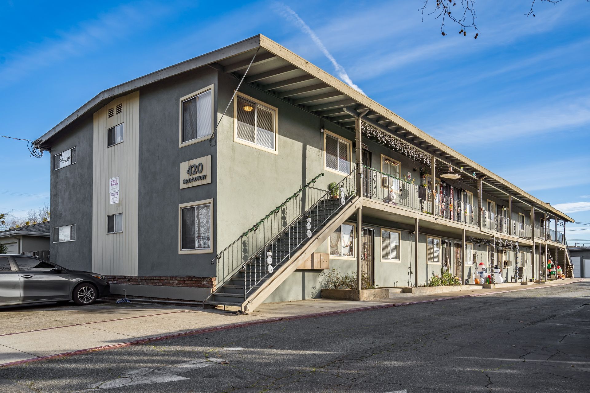 Gray two-story apartment building with stairwell and parked car on a sunny day.