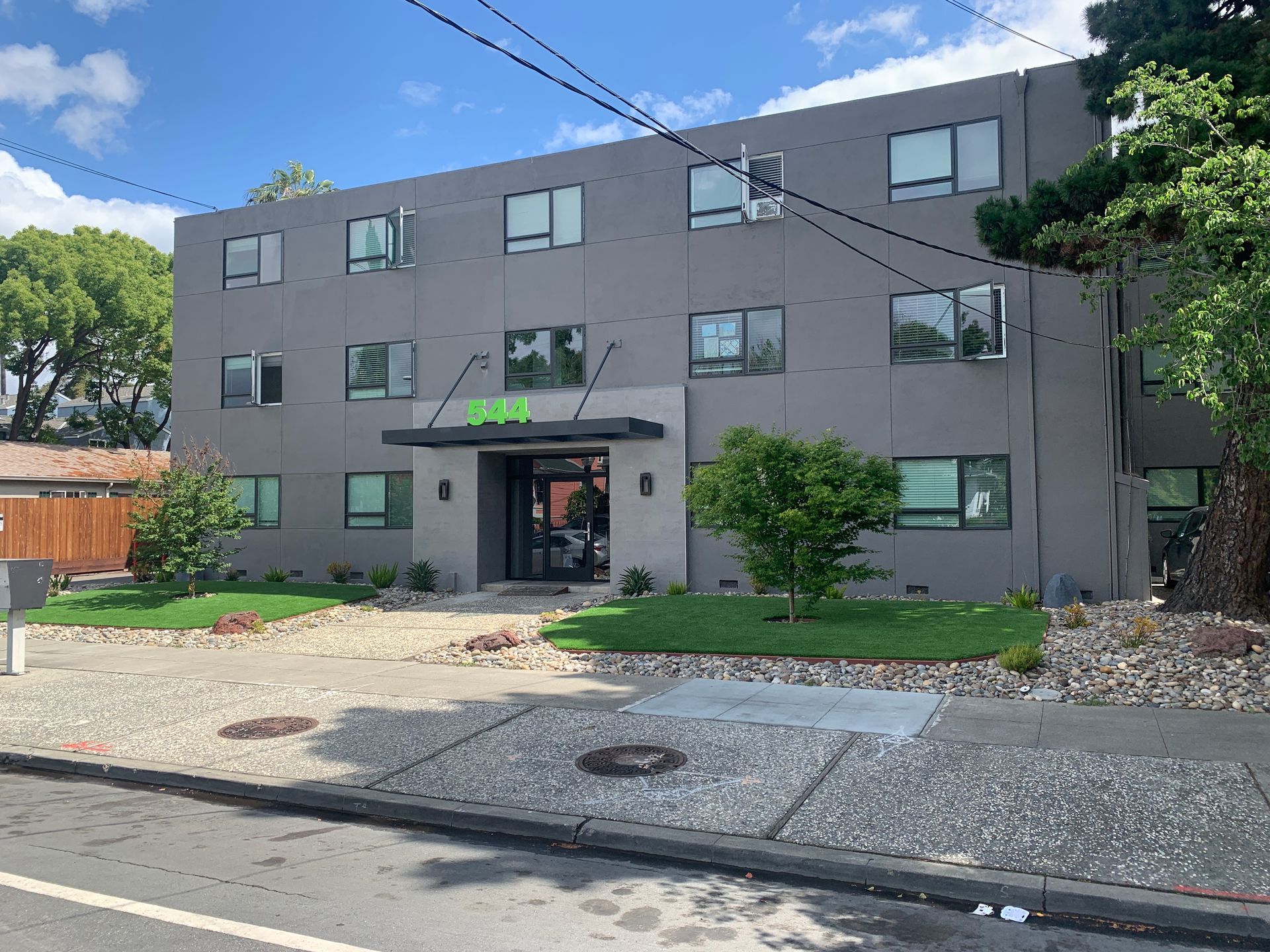 Two-story apartment building with brick and beige facade, shaded by trees, with a driveway in front.