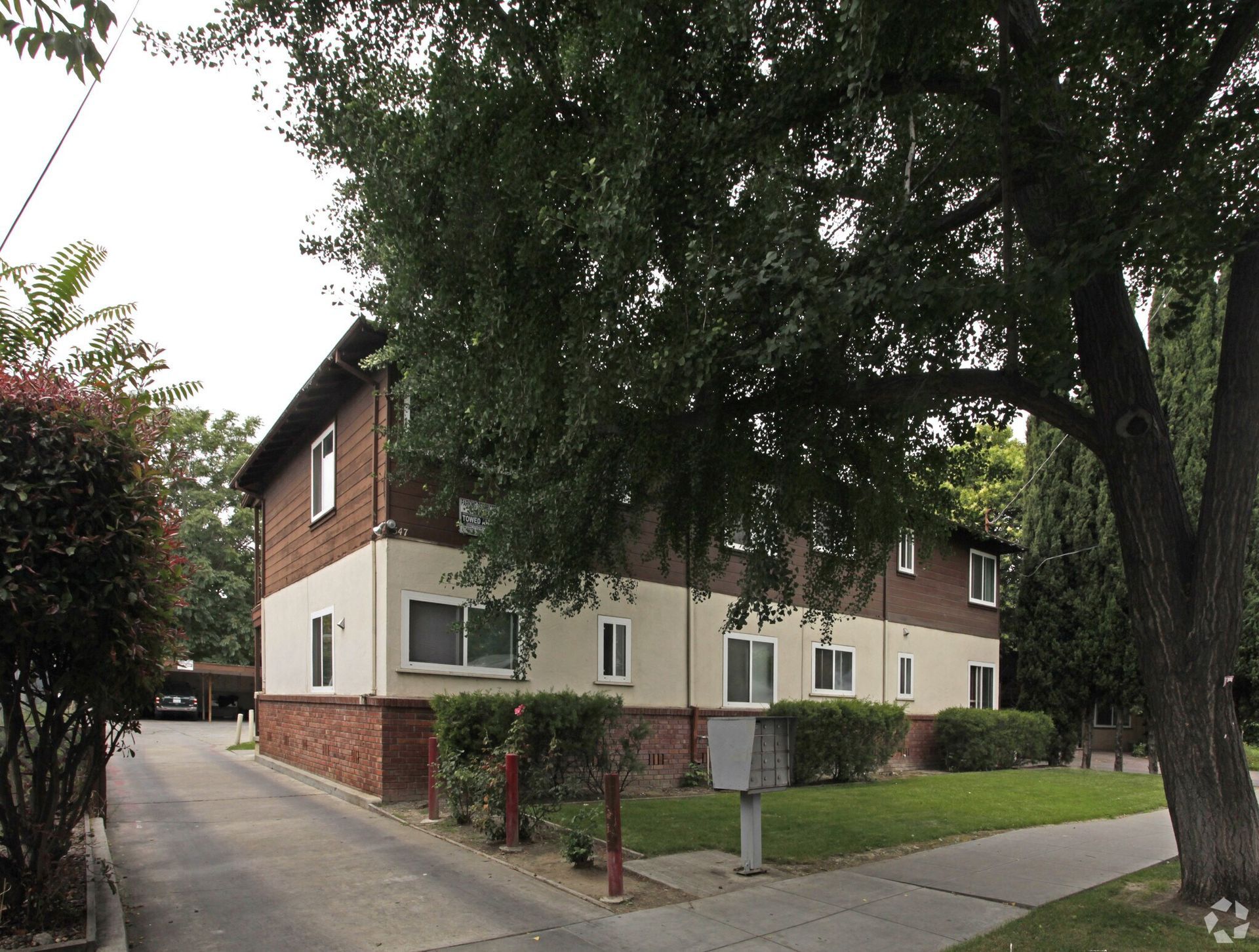 Two-story apartment building with brick base, tan walls, and brown top story, partially obscured by trees.