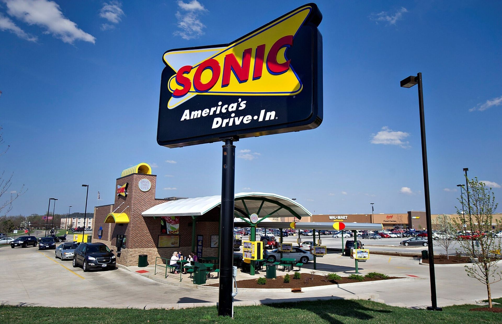Sonic Drive-In restaurant with a large sign, drive-up stalls, and cars. Blue sky background.