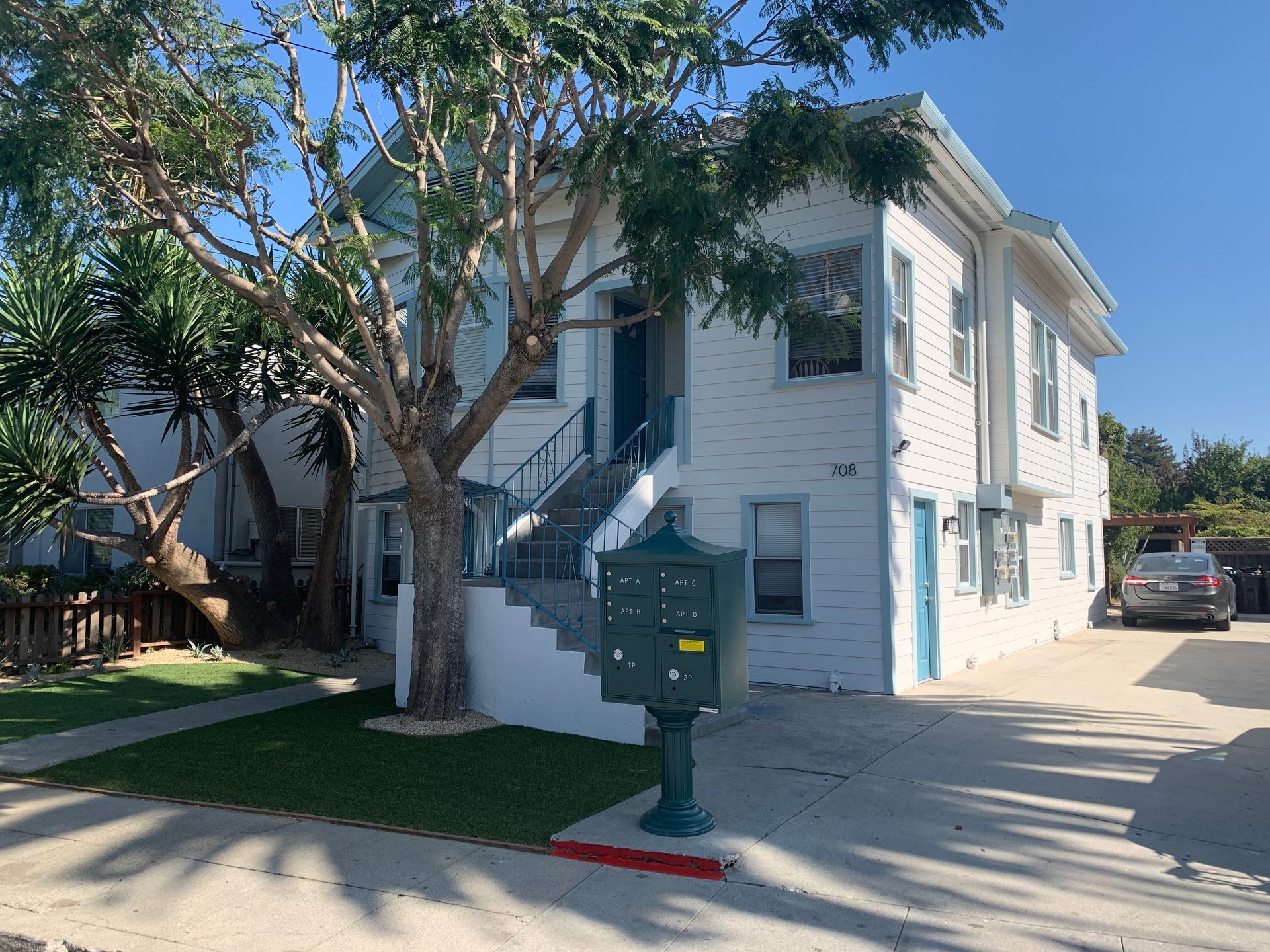 Two-story white building with blue accents, mailbox, tree, and parked car on a sunny day.