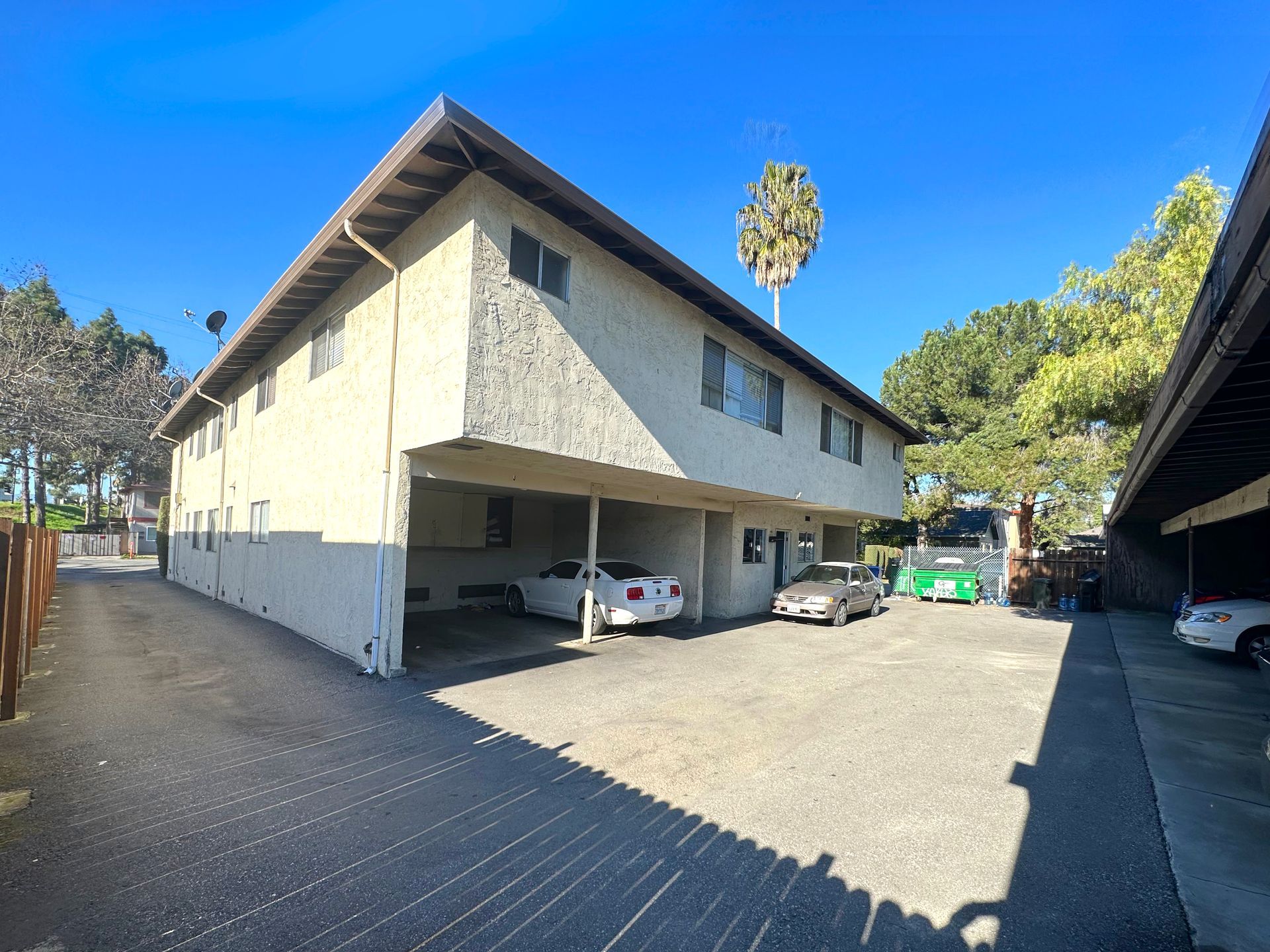 Two-story apartment building with covered parking, driveway, and palm tree under a blue sky.
