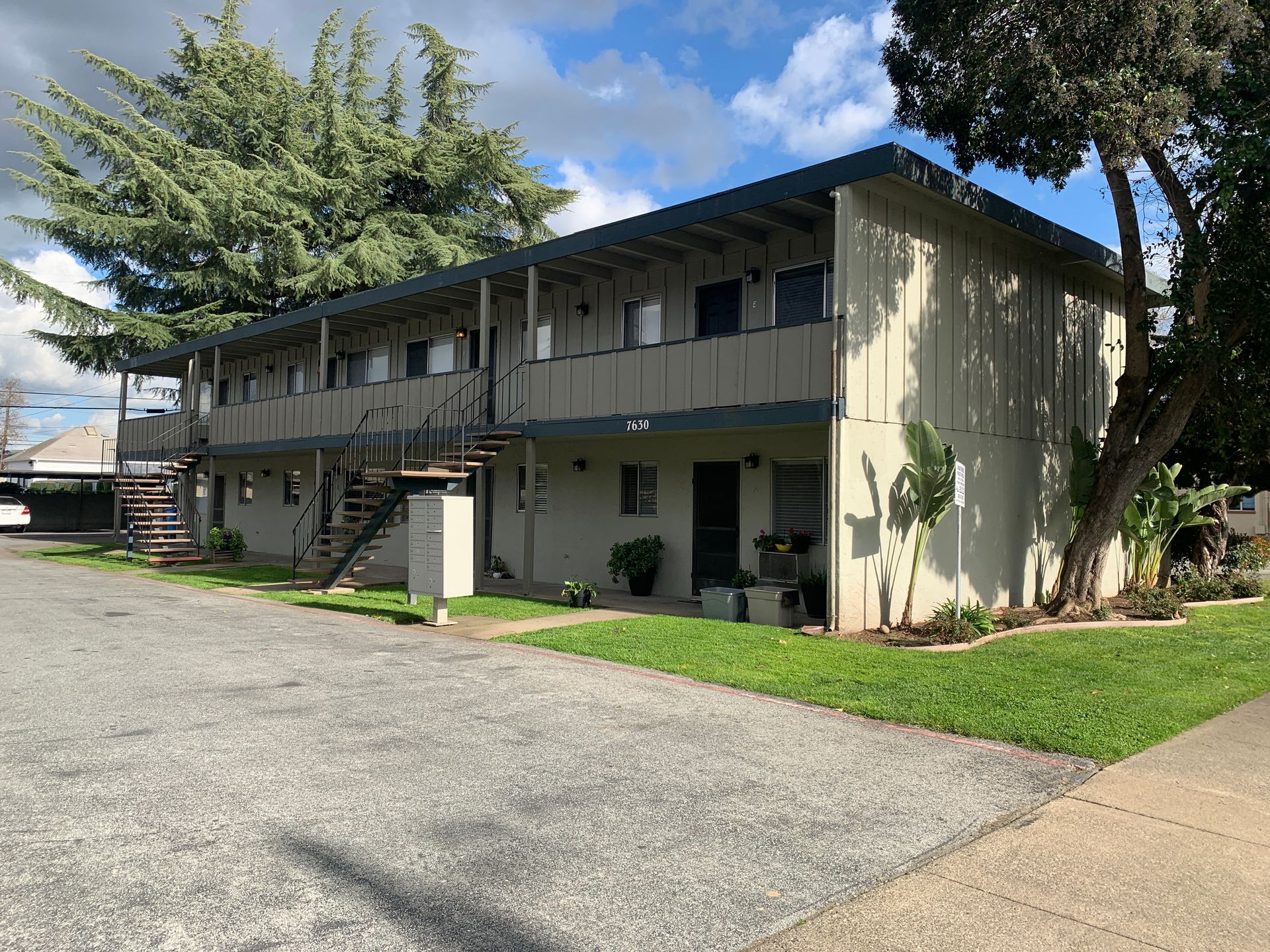 Two-story beige apartment building with external staircases, a blue roof, and a gravel parking lot.