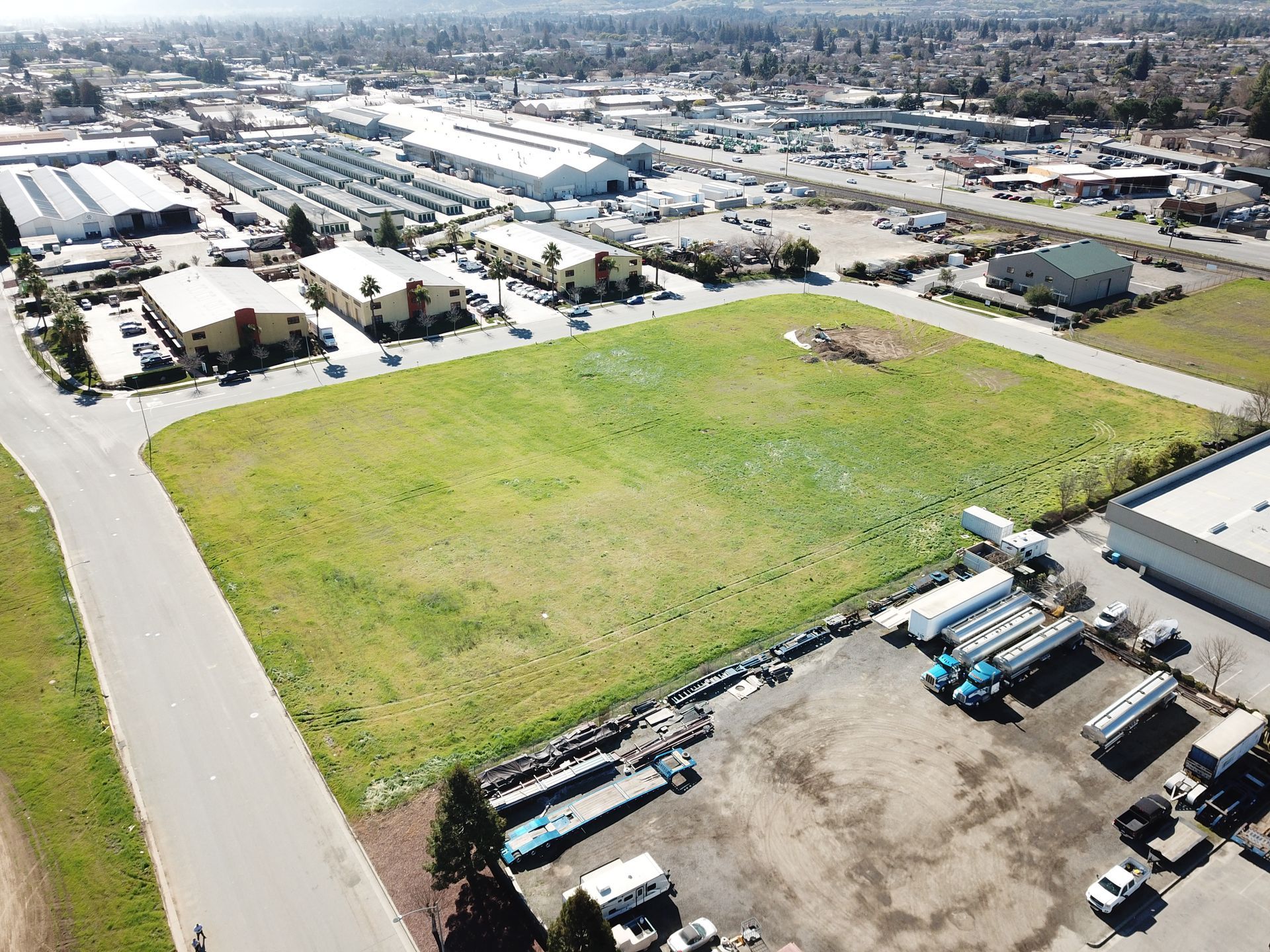 Aerial view of a vacant, grassy lot surrounded by industrial buildings and a road; sunny day.