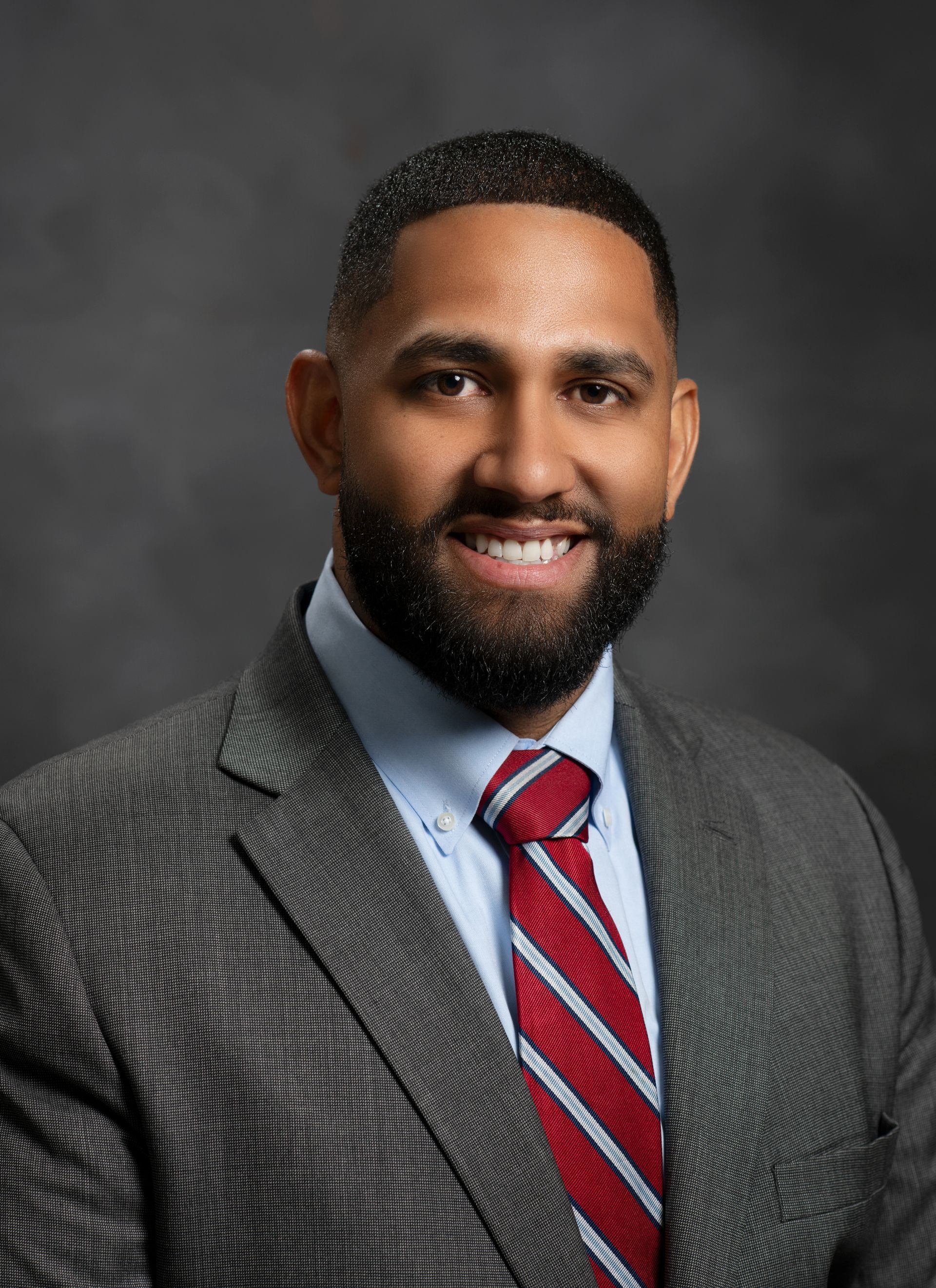 Man in gray suit and blue shirt smiles, headshot against a gray backdrop.