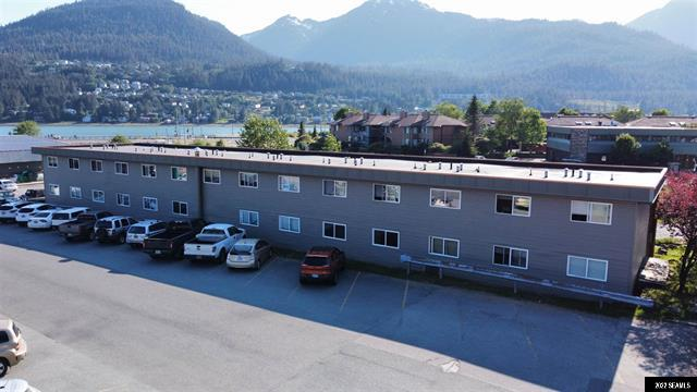 Gray two-story building with cars parked in front; mountains and water in the background.