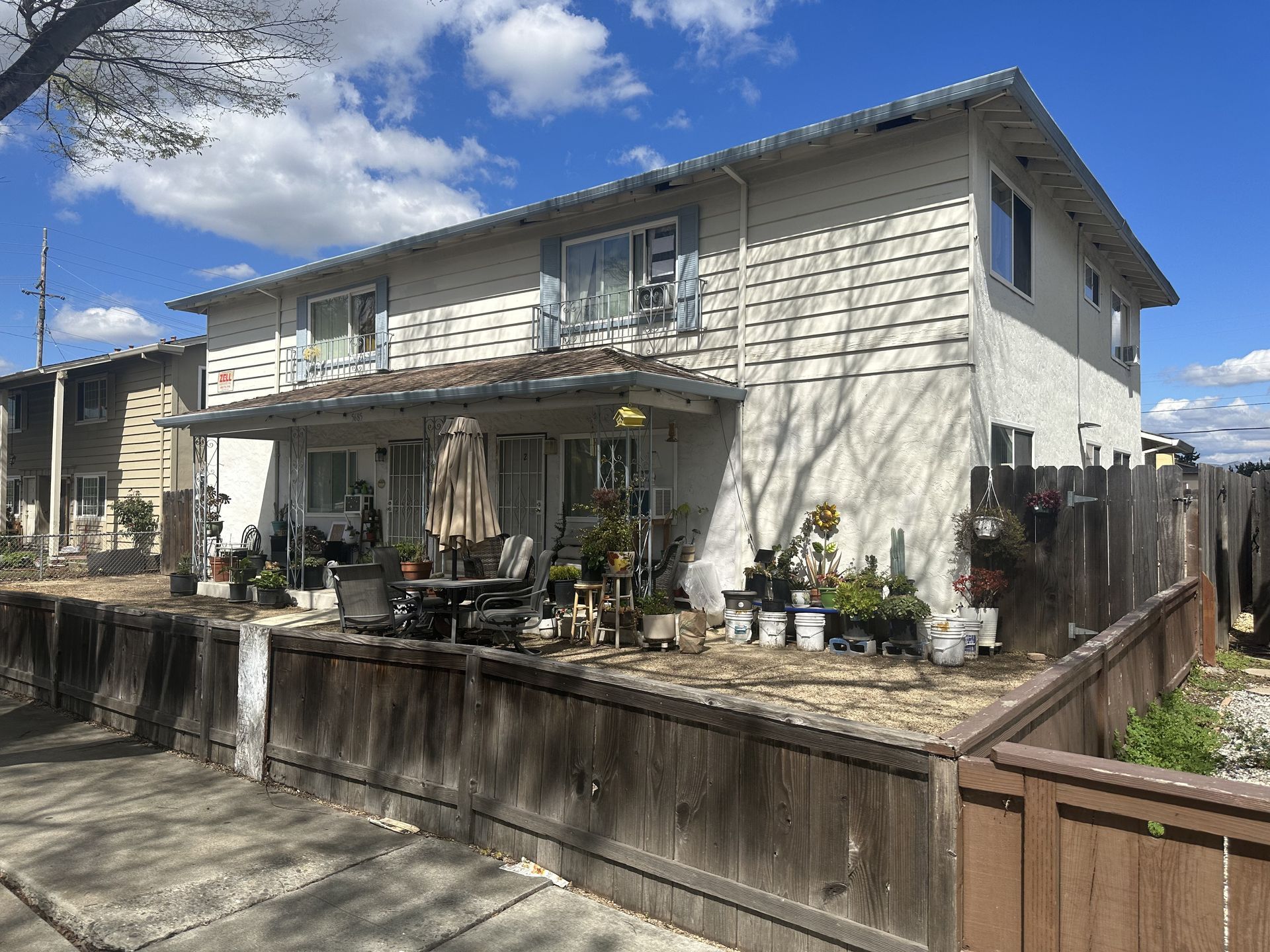 Two-story tan apartment building with a wooden fence and various potted plants in front.