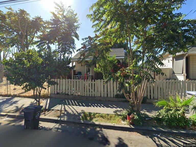 Small house behind a white picket fence, trees, and trash bin along the street.