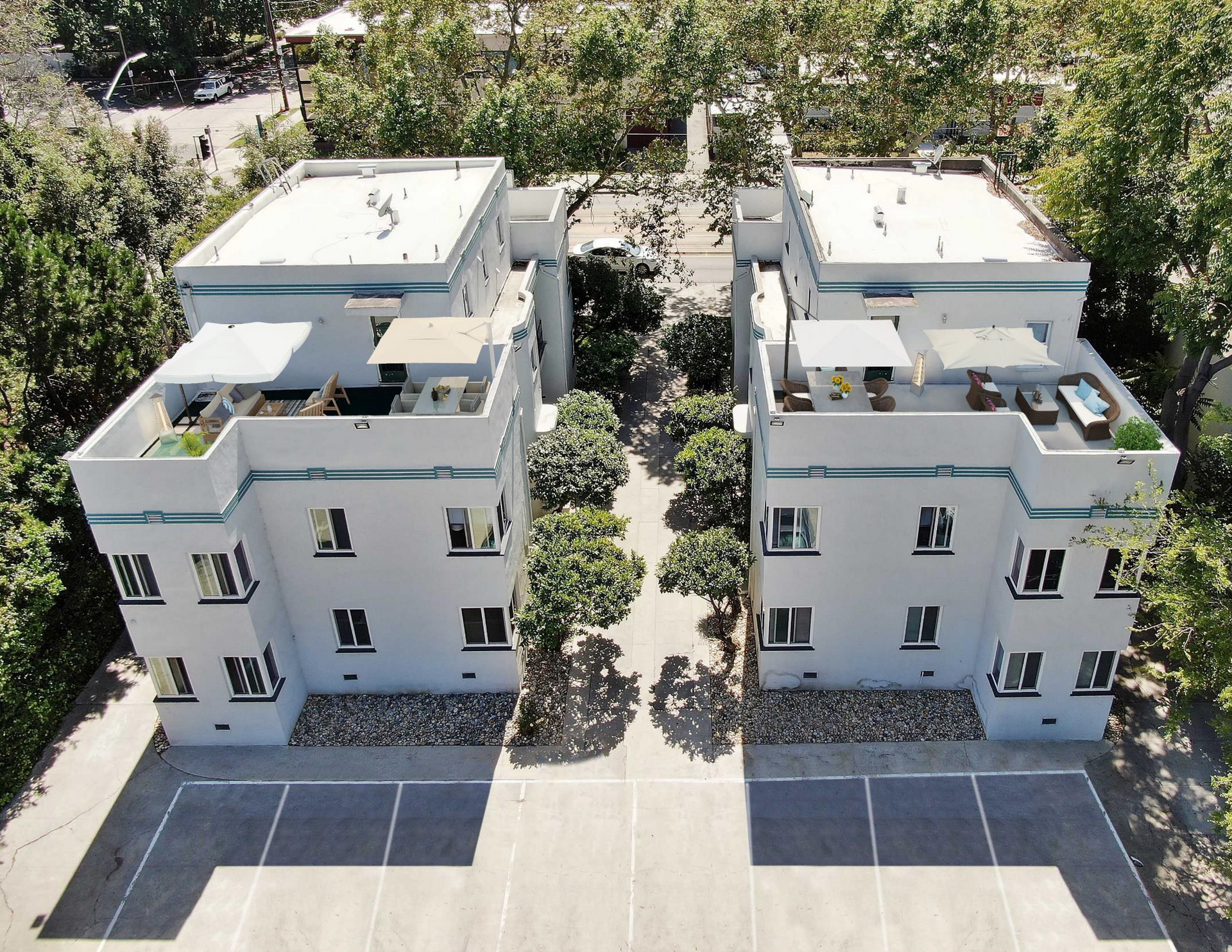 Aerial view of two white, three-story apartment buildings with flat roofs, connected by a path of trees.