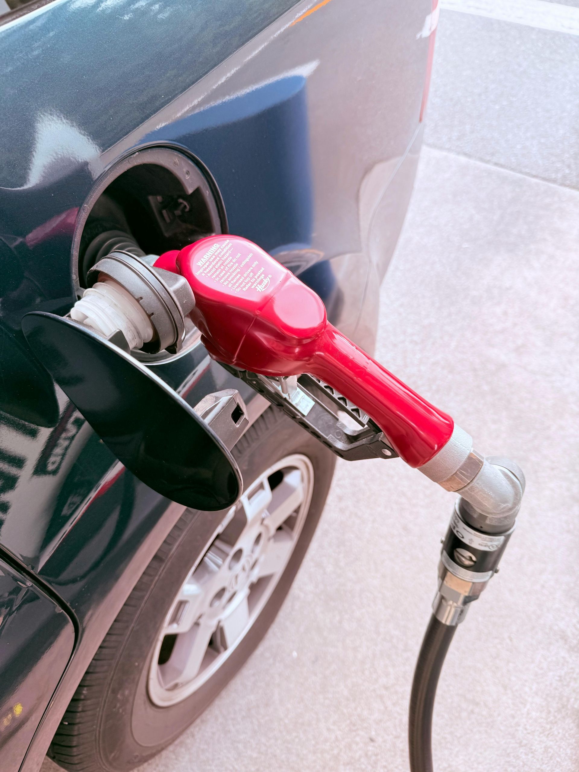 Red gas pump nozzle filling a dark blue car's fuel tank.