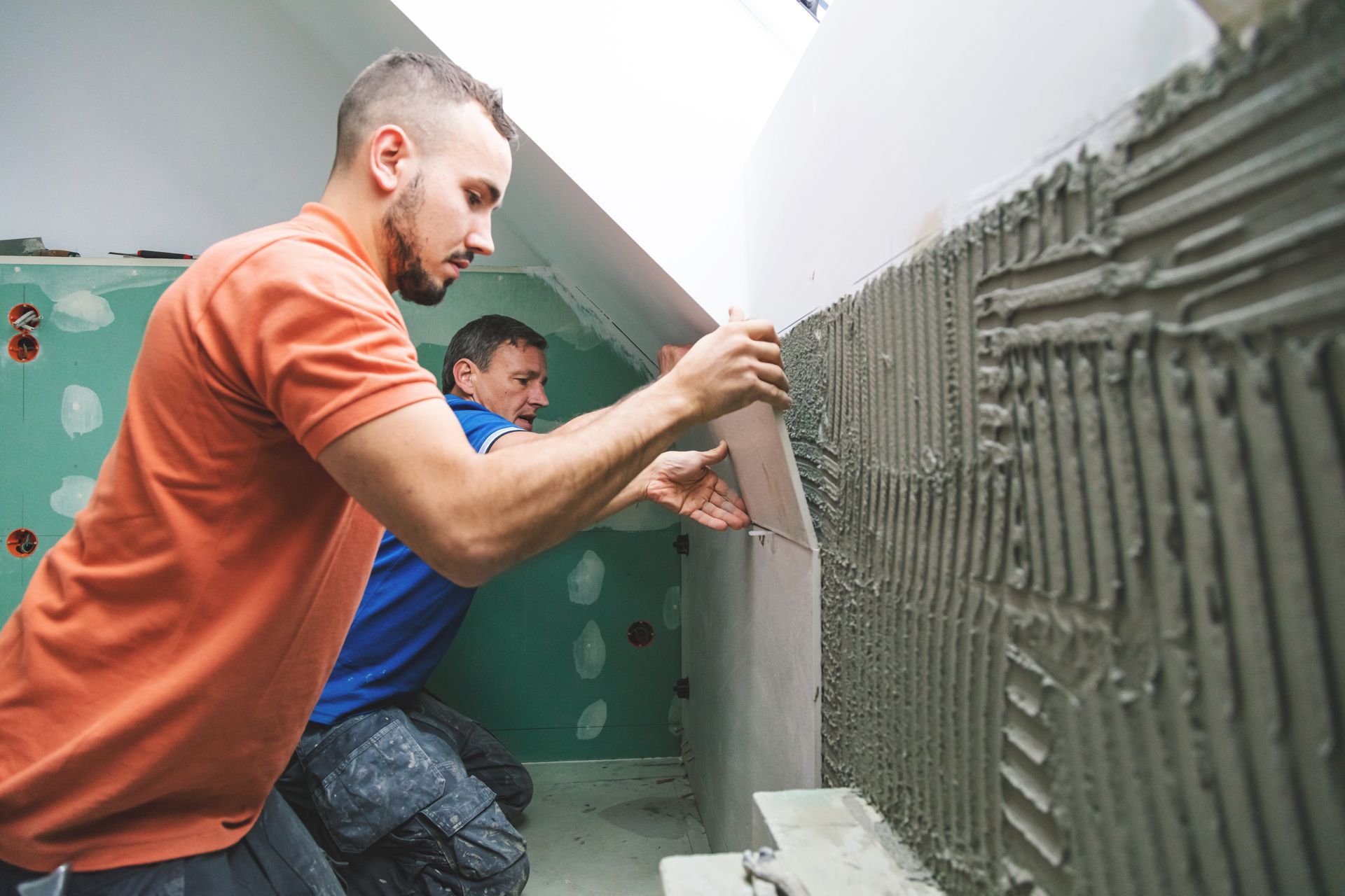 Two people tiling a wall, applying a tile to a mortar-covered surface.