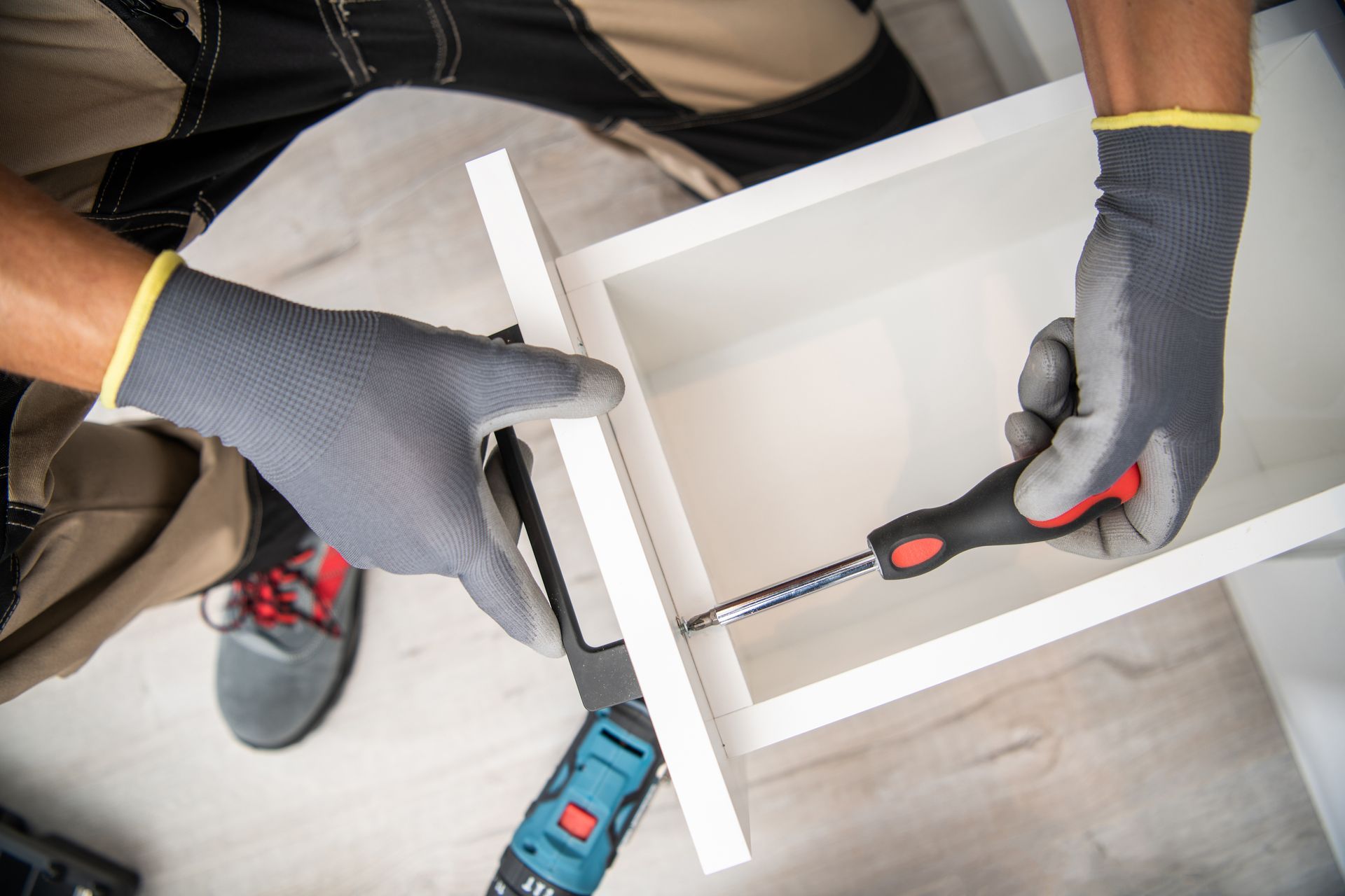Person assembling white furniture with a screwdriver, wearing work gloves.
