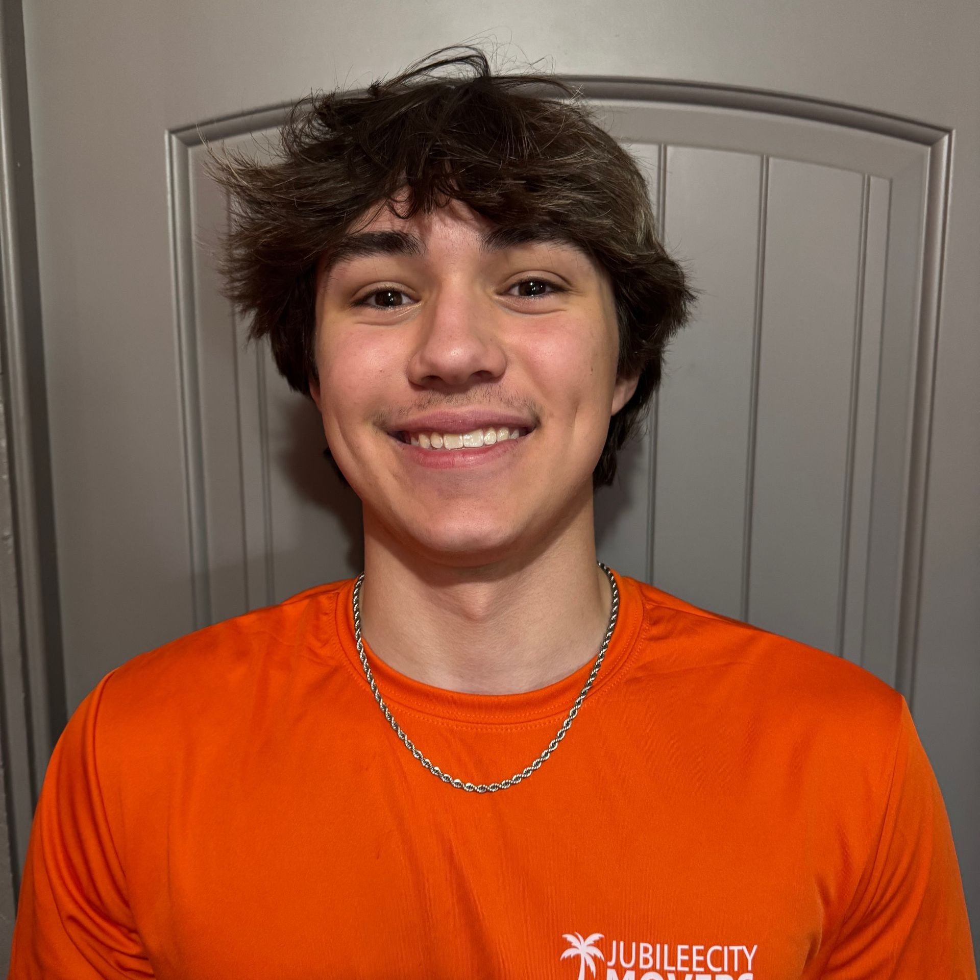 A young man wearing a black shirt and a gold chain is smiling in front of an orange wall.