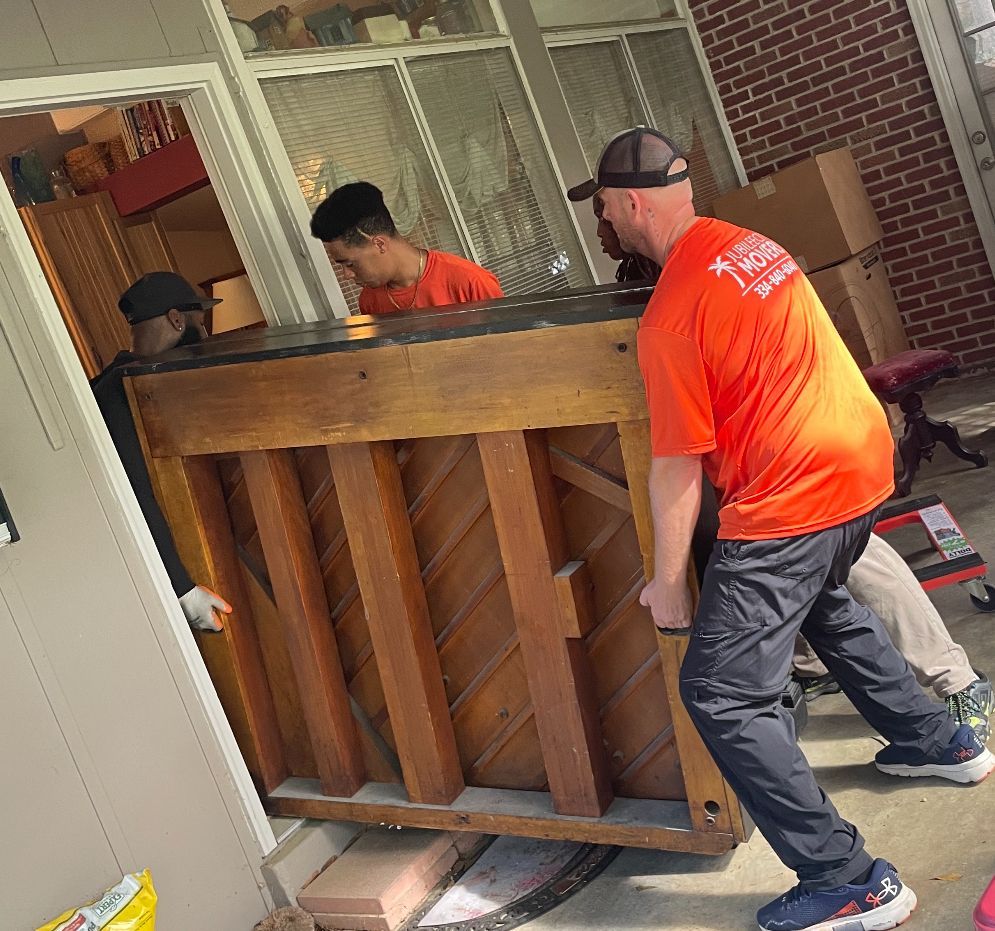 A man in an orange shirt is carrying a piano on a cart.