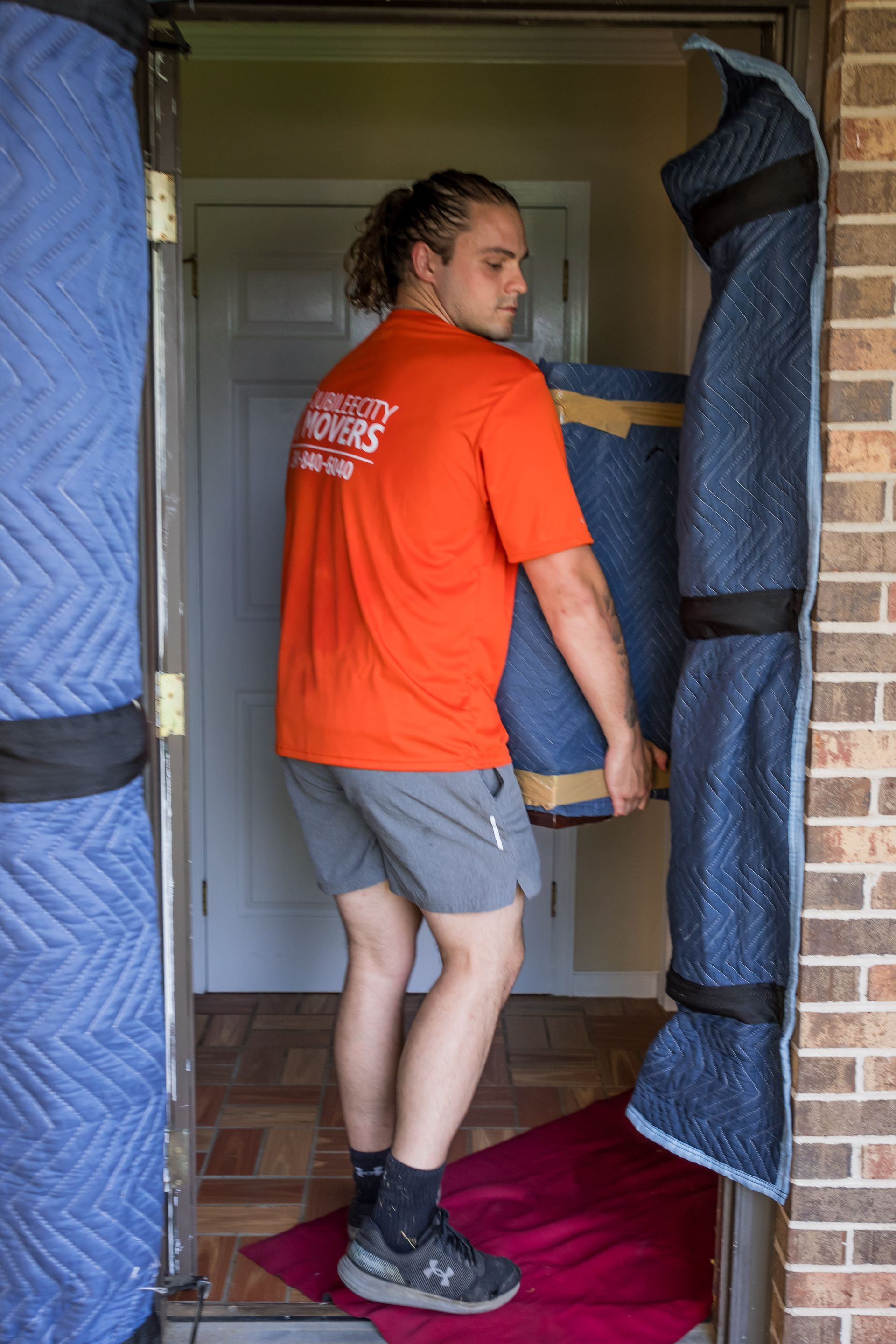 A man in an orange shirt is carrying a blue couch in a doorway.