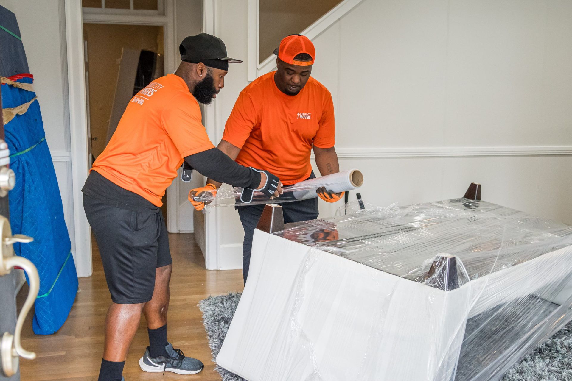 Two men are wrapping a table with plastic wrap in a living room.
