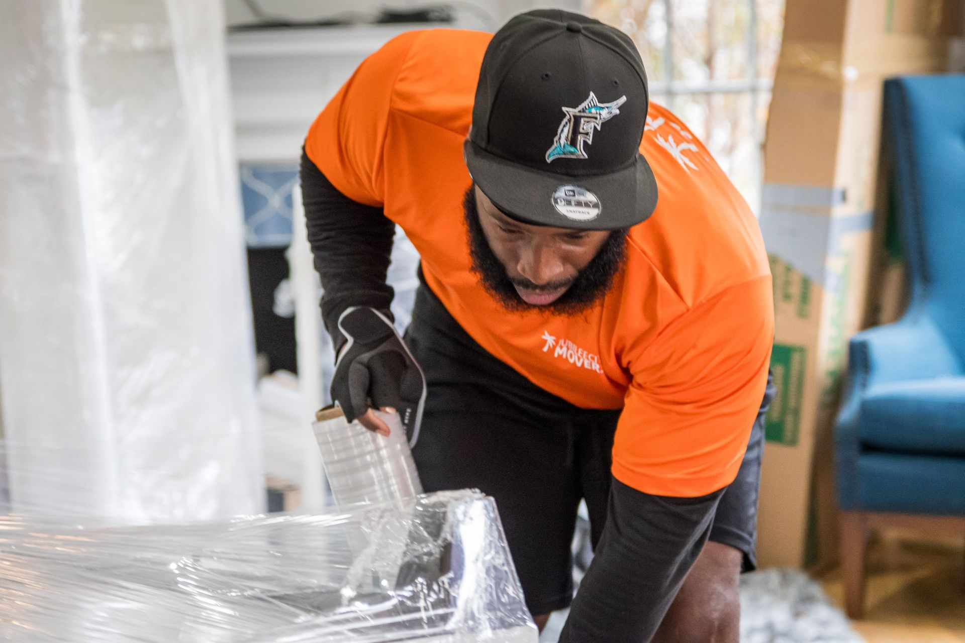 A man in an orange shirt and black hat is wrapping something in plastic.